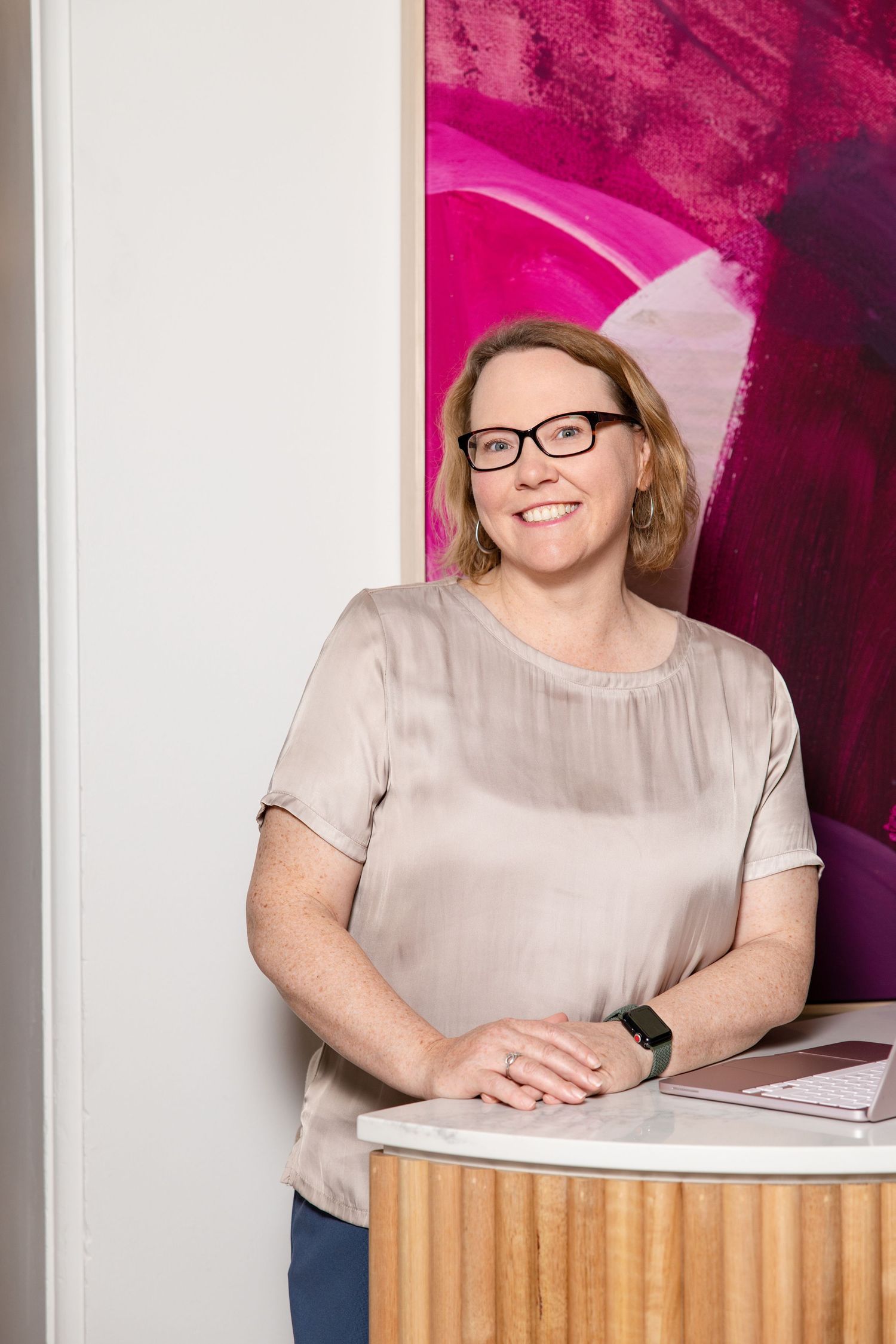 Smiling woman with glasses in a beige blouse leaning on a wooden desk with pink abstract art behind.
