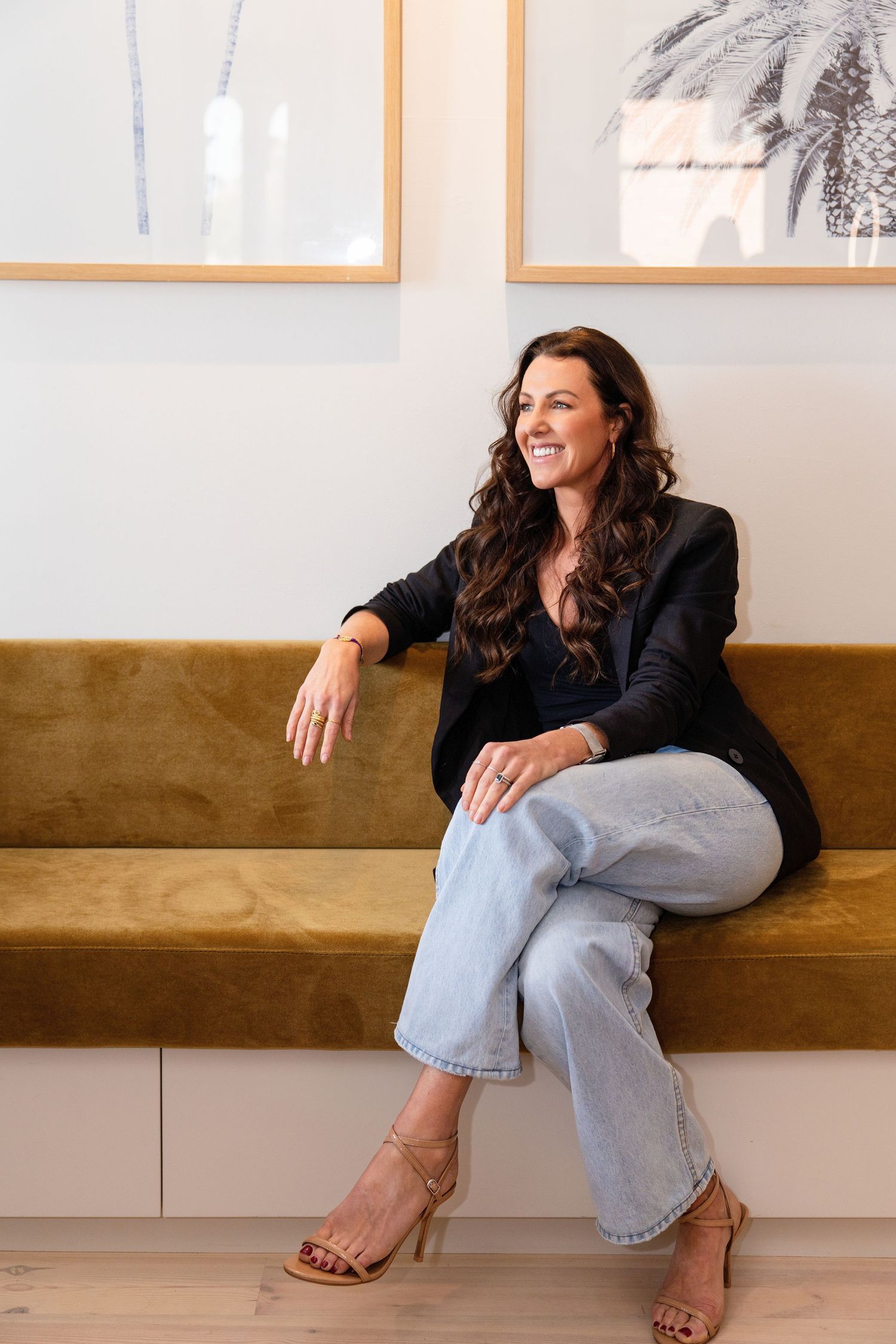 Brunette woman in black blazer and jeans smiling, seated on gold velvet bench with framed art behind her.