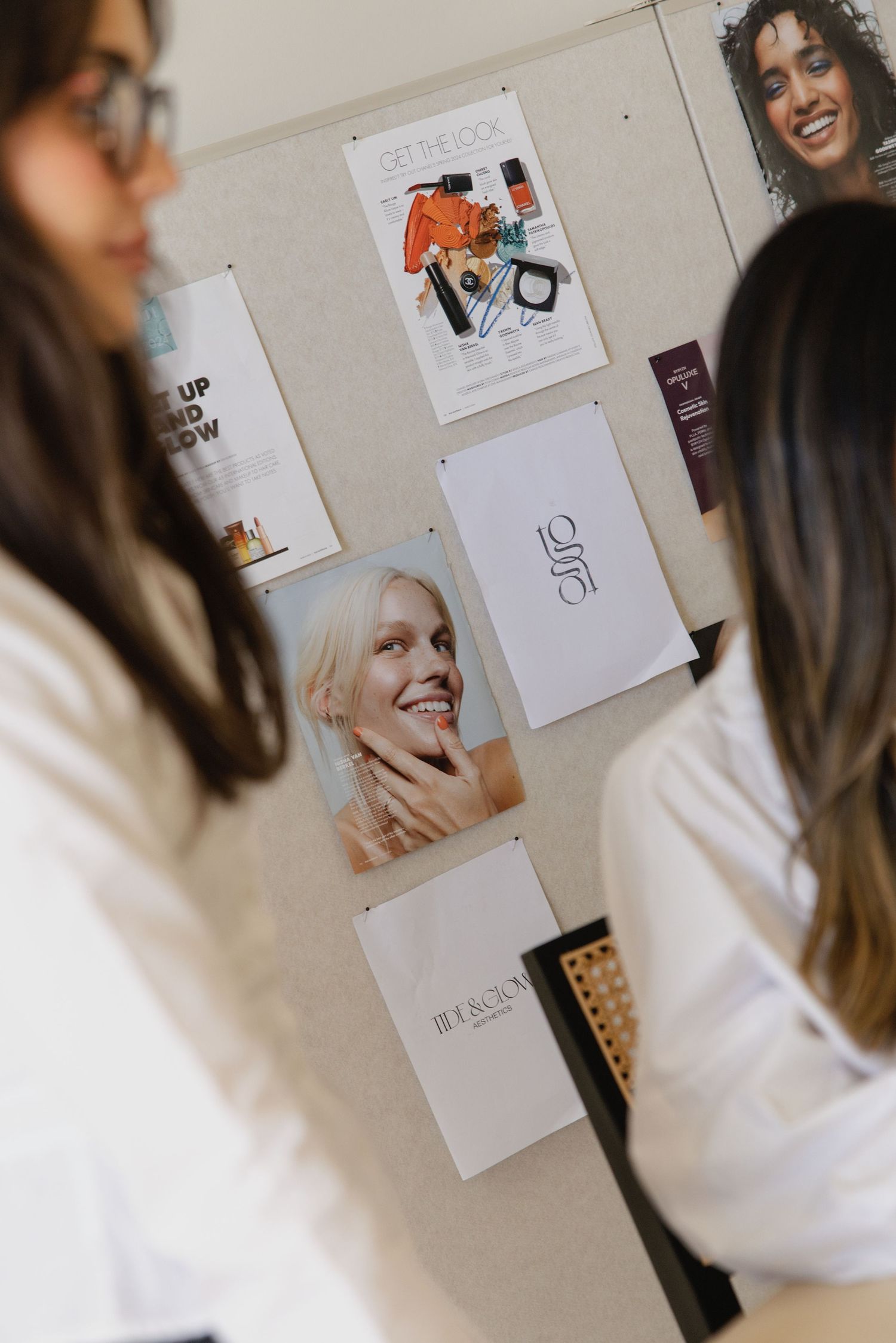 Two women in white shirts reviewing beauty brand mood board with cosmetic posters and logos pinned to wall.