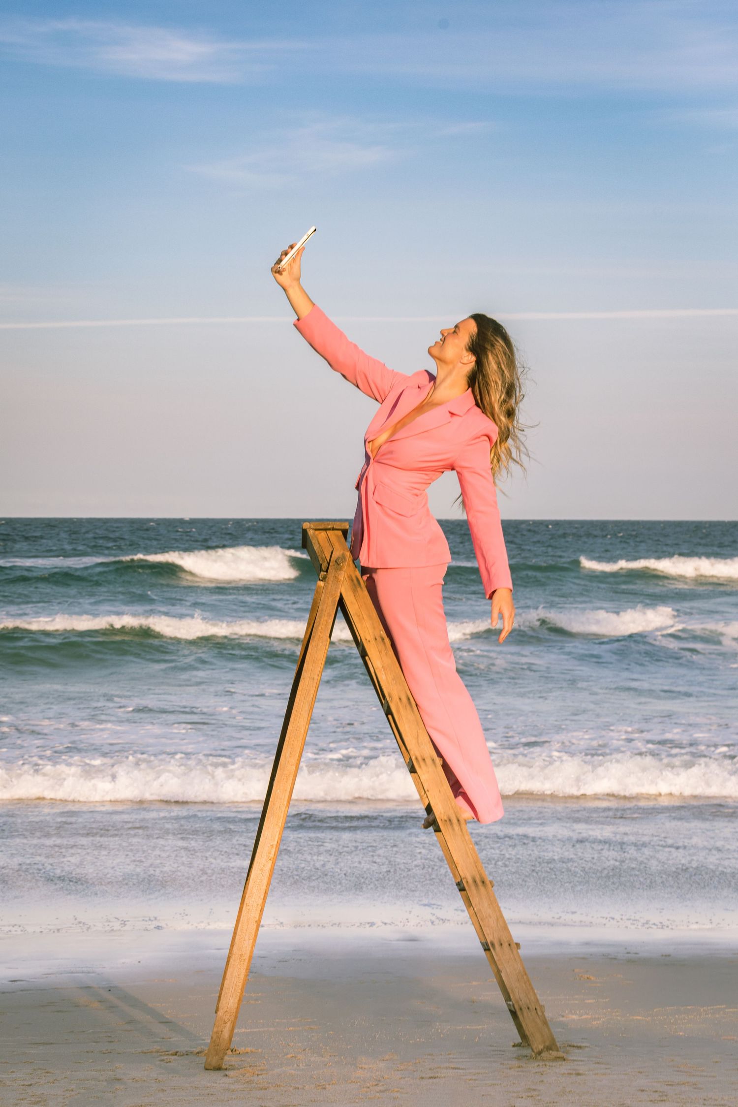 A person in pink clothing strikes dynamic poses against a wooden post on a sunny beach with ocean waves in the background.