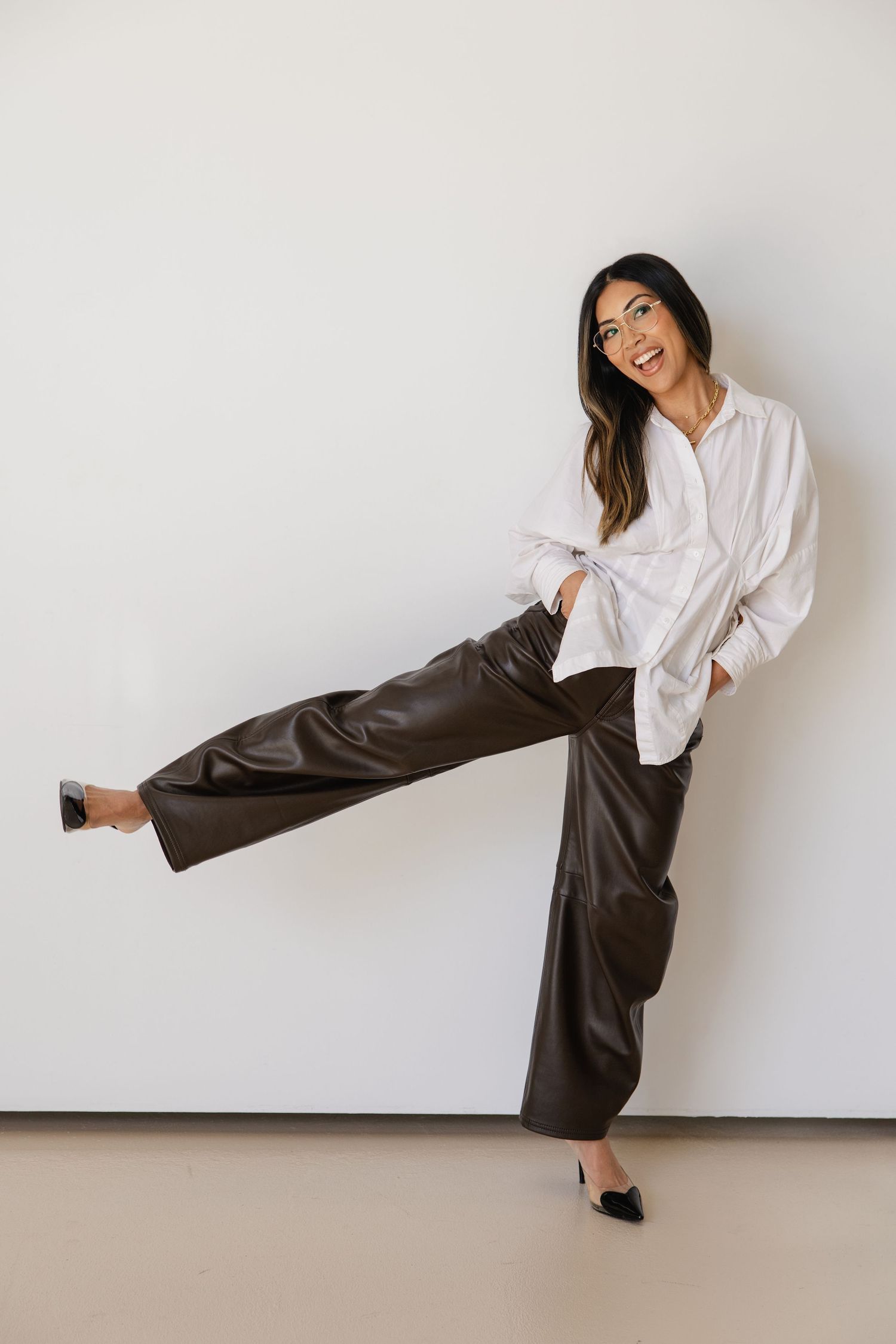 Woman in white shirt and dark brown wide-leg leather pants kicking leg up, smiling against white wall.
