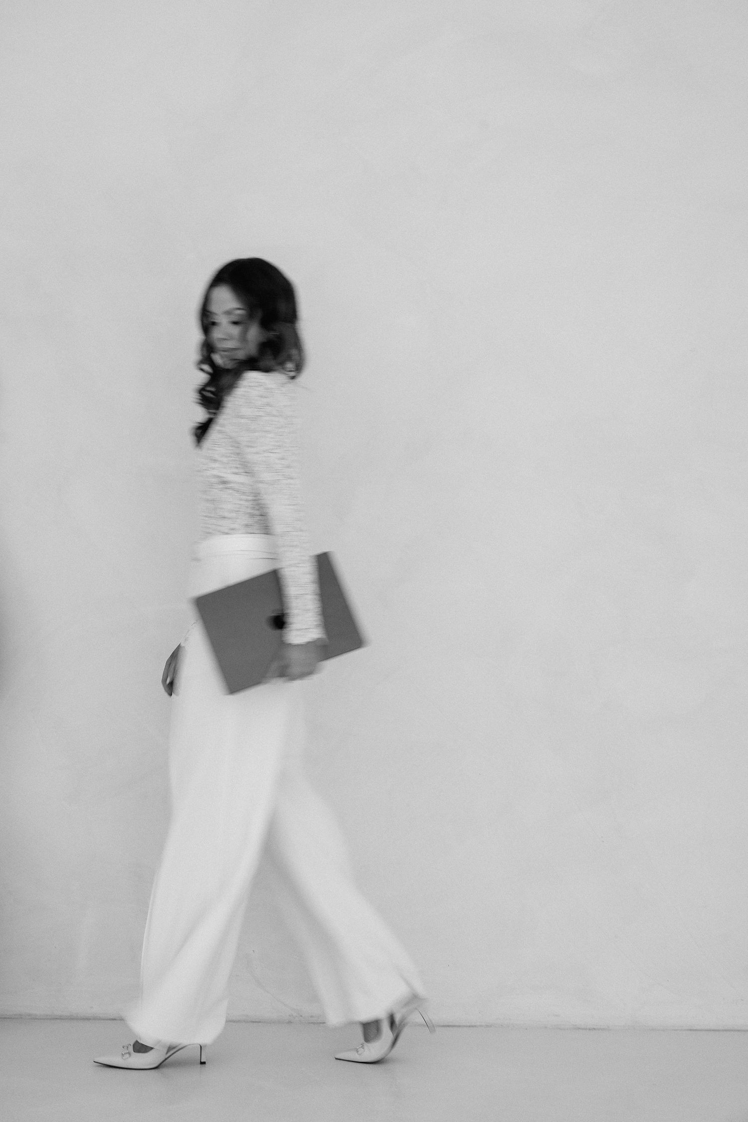 Black and white photo of a woman in white wide-leg pants and striped top carrying a laptop while walking.