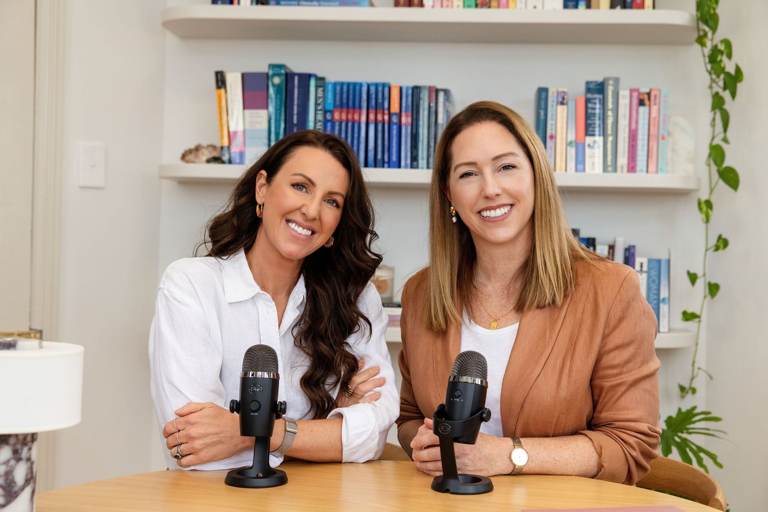 Two women sitting at a desk with microphones, smiling in a cozy home office setting.