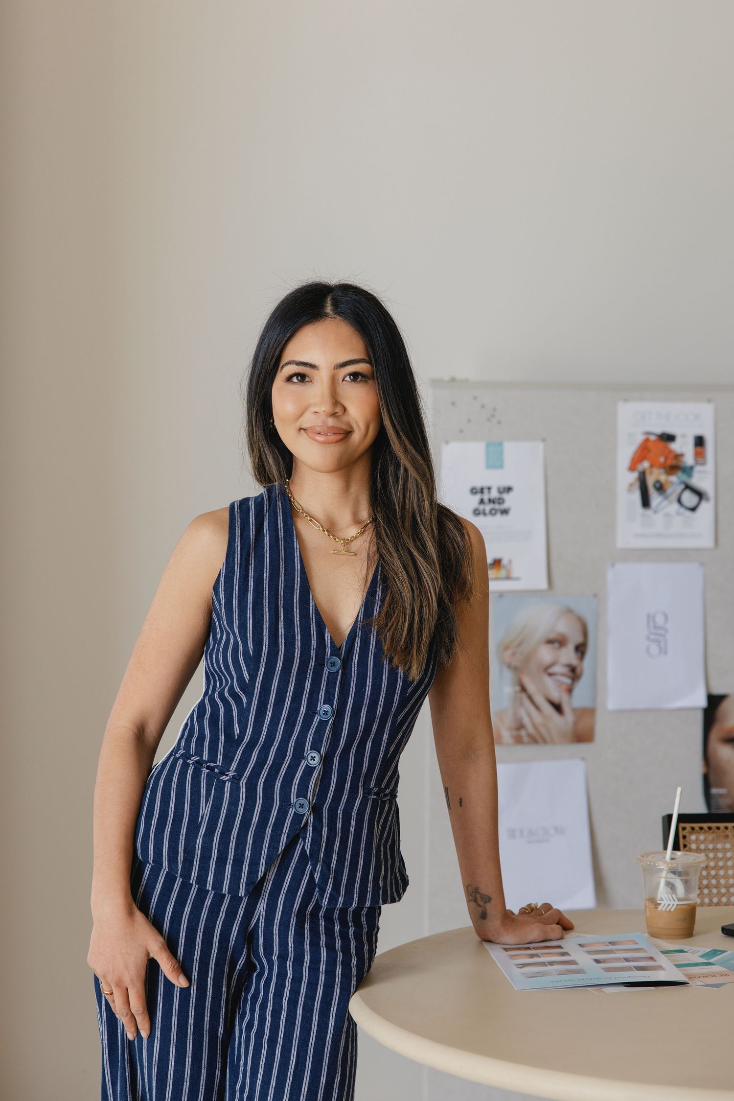 Woman in navy striped vest and pants leaning against a desk in a modern office workspace with mood board in background.