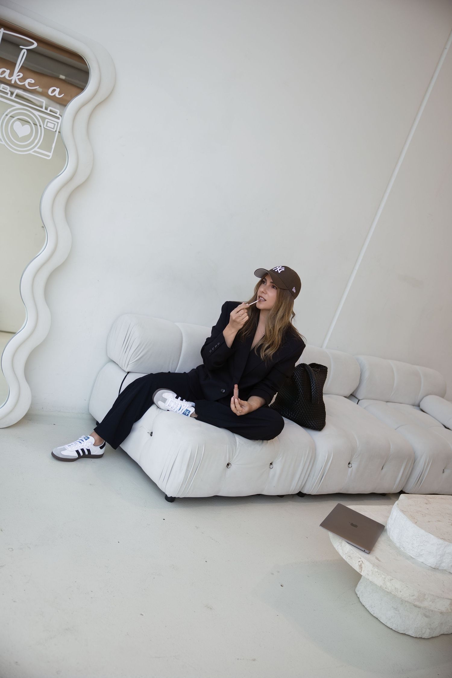 Woman in black suit and cap sitting on white sofa in minimalist white room.