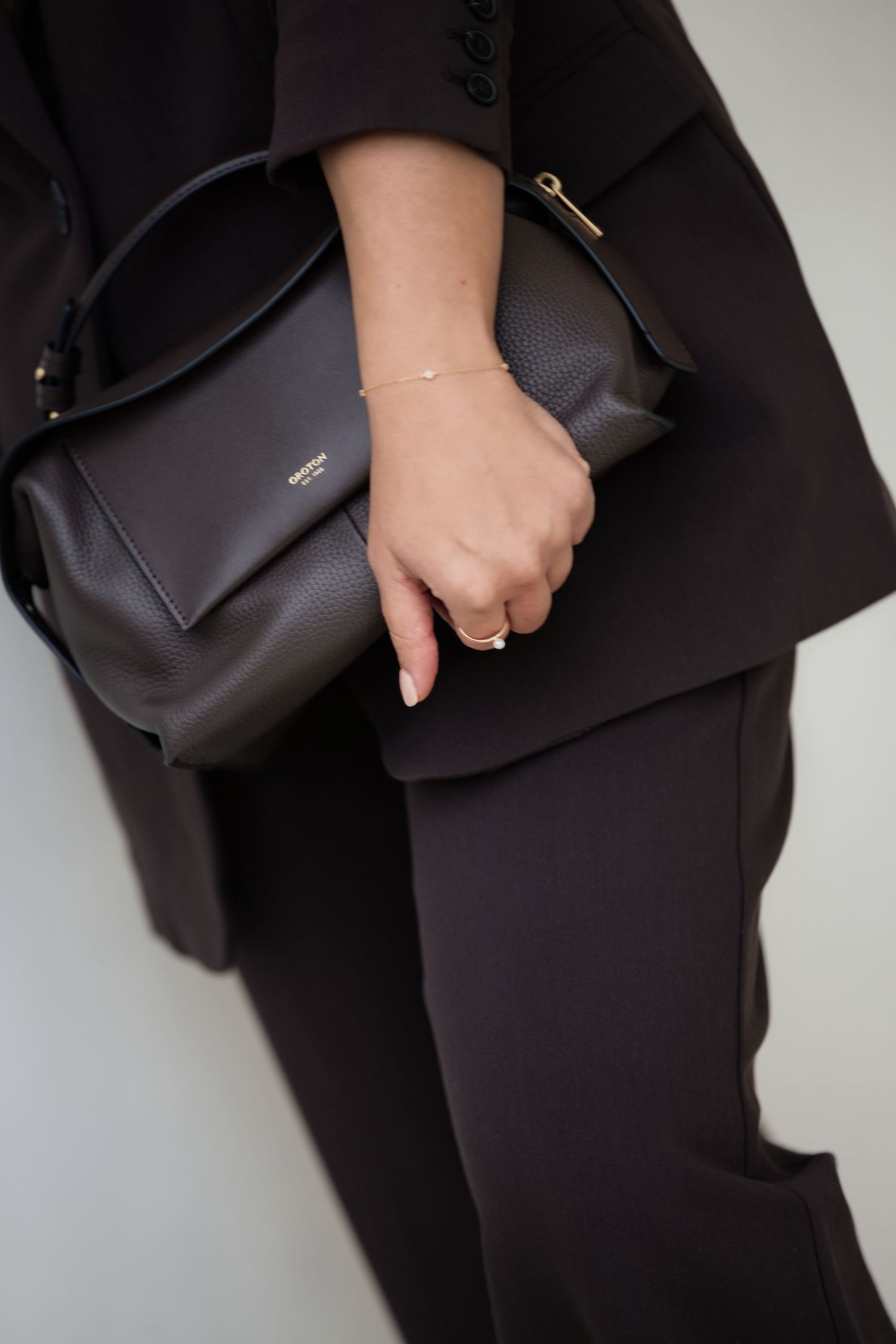 Woman in black suit holding a dark brown leather handbag with gold hardware and delicate bracelet.