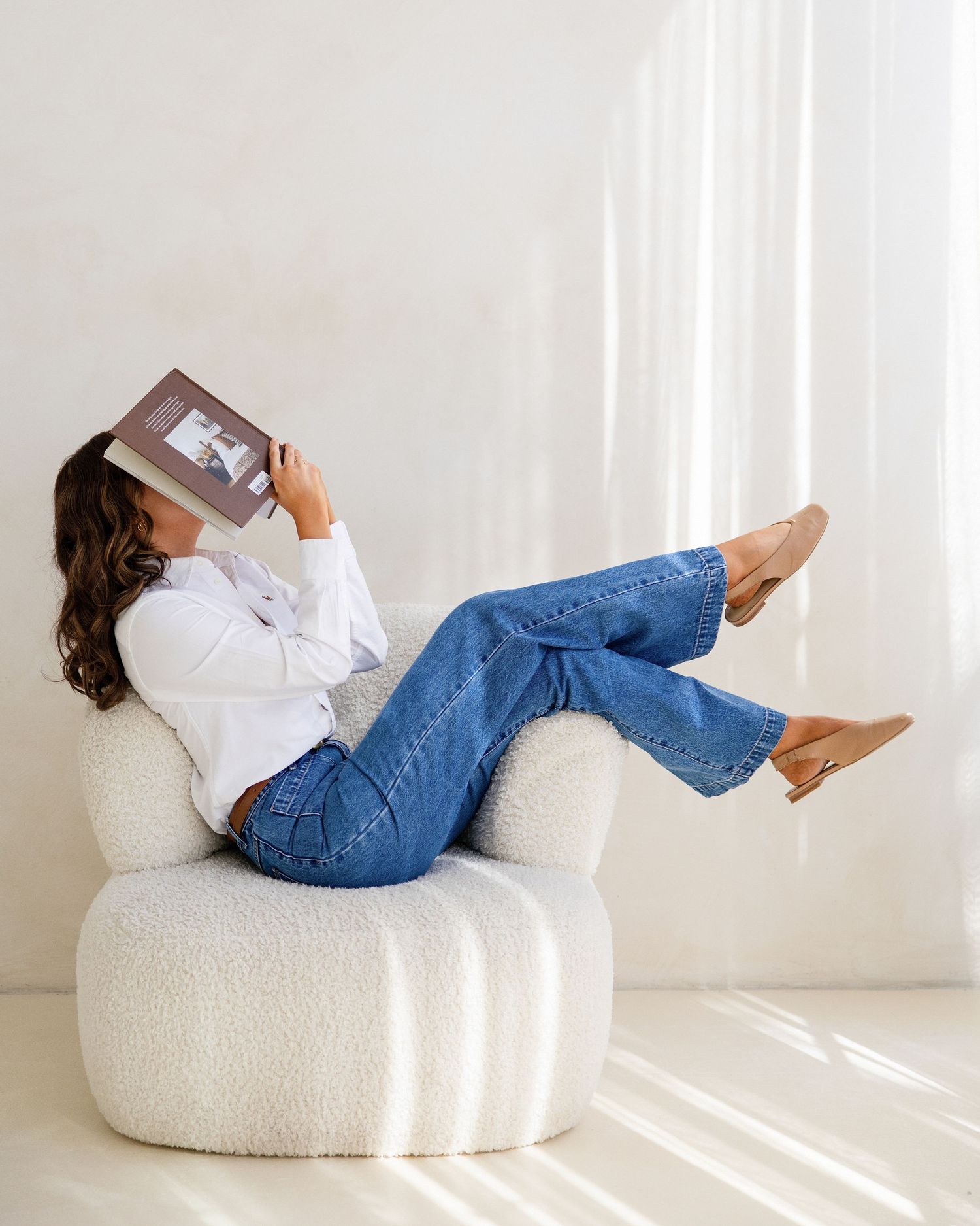 Woman relaxing in white chair reading a book, wearing white shirt and blue jeans.