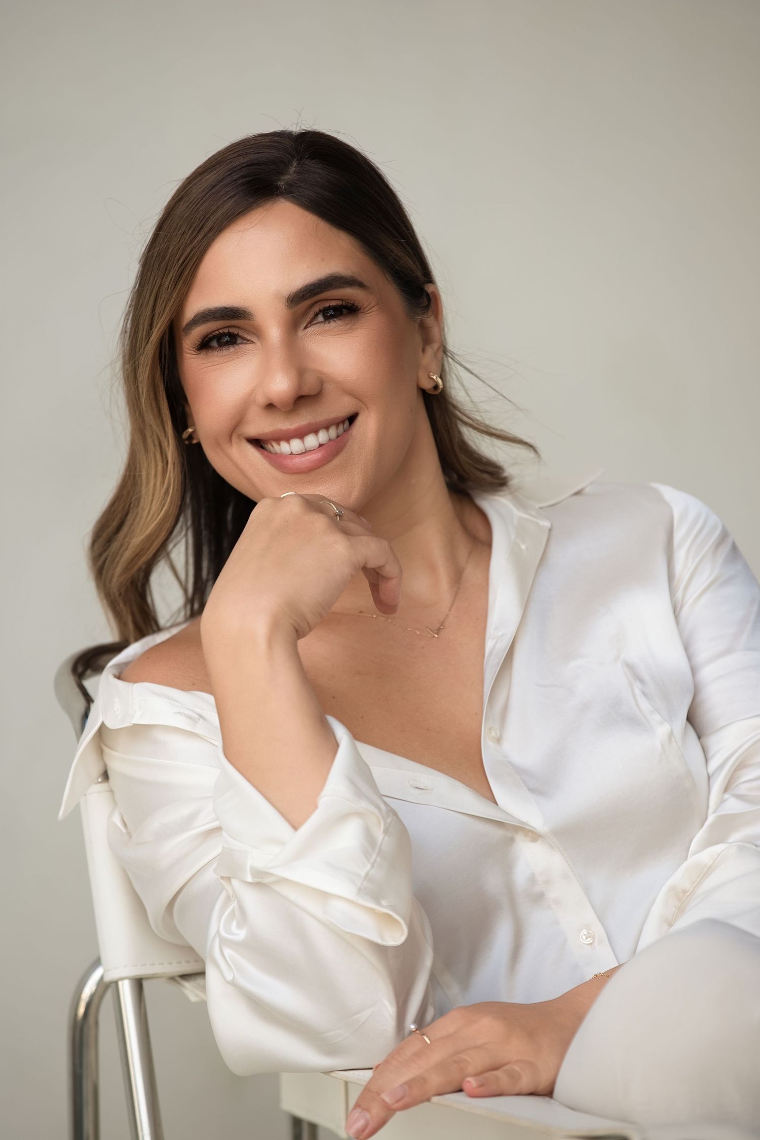 Smiling woman in white satin blouse sitting on chair with hand on chin against light background.