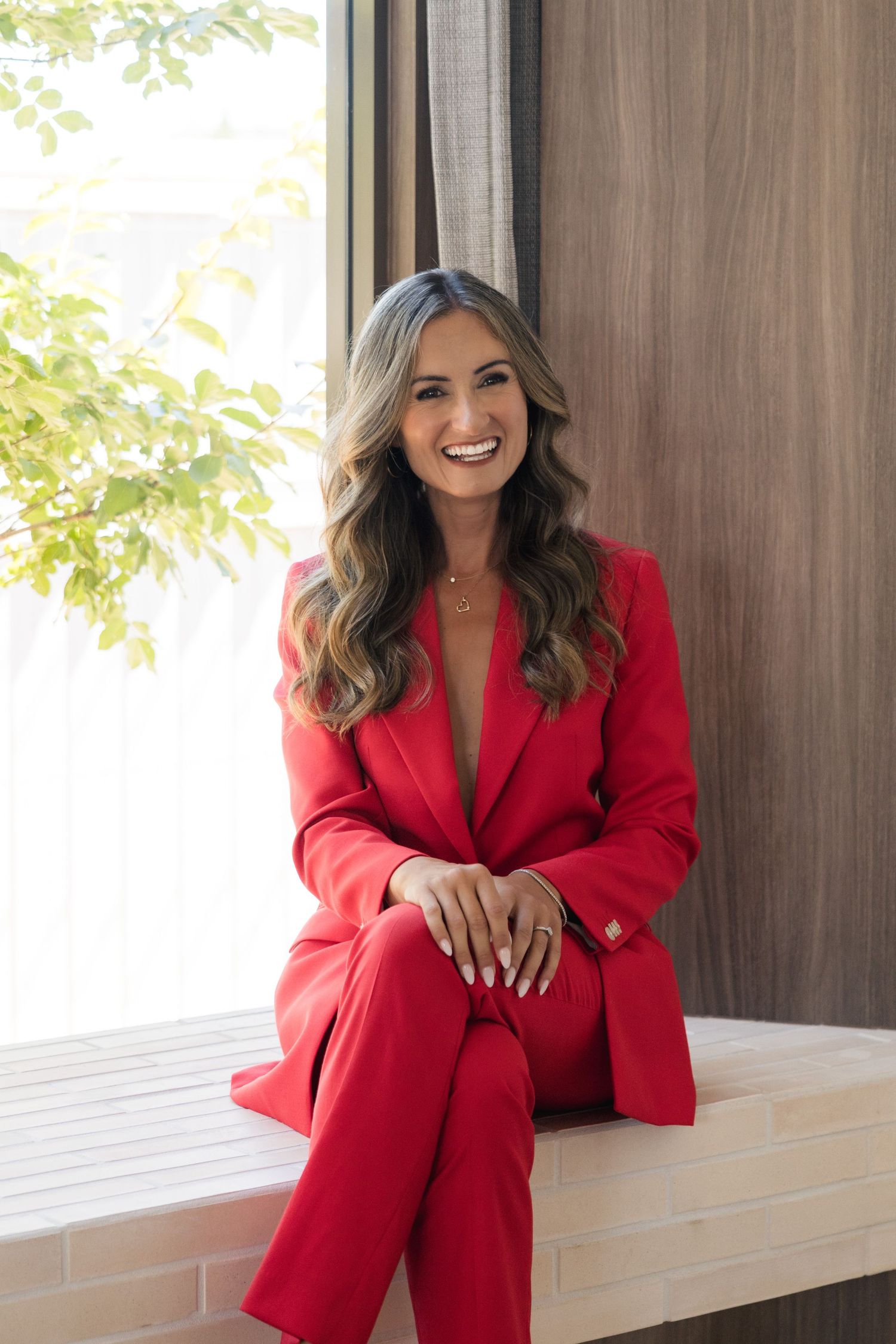 Confident woman in red pantsuit sitting by window with green foliage background, smiling warmly.