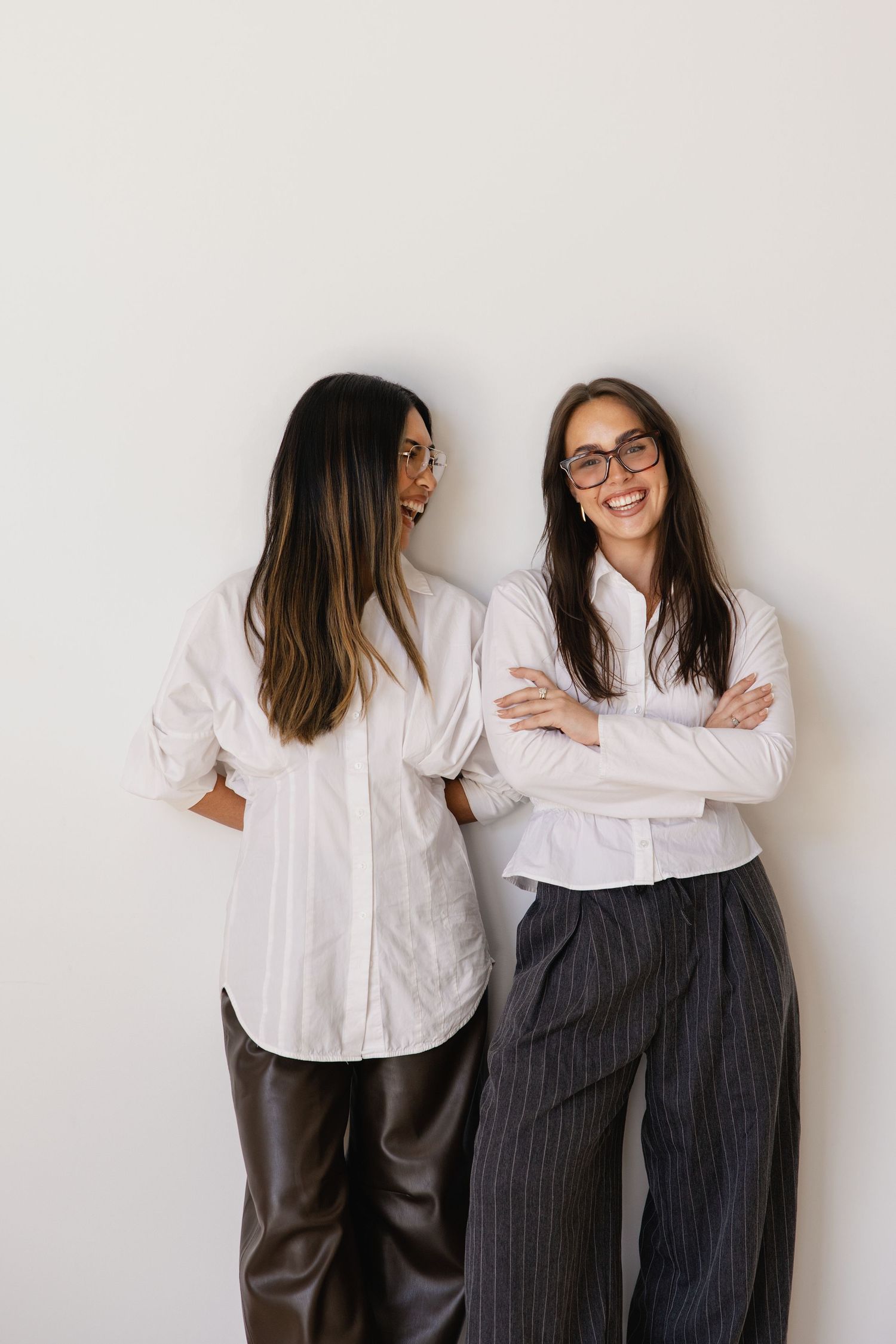 Two women in white shirts and glasses smile together against a white wall, one in leather pants, one in striped trousers.