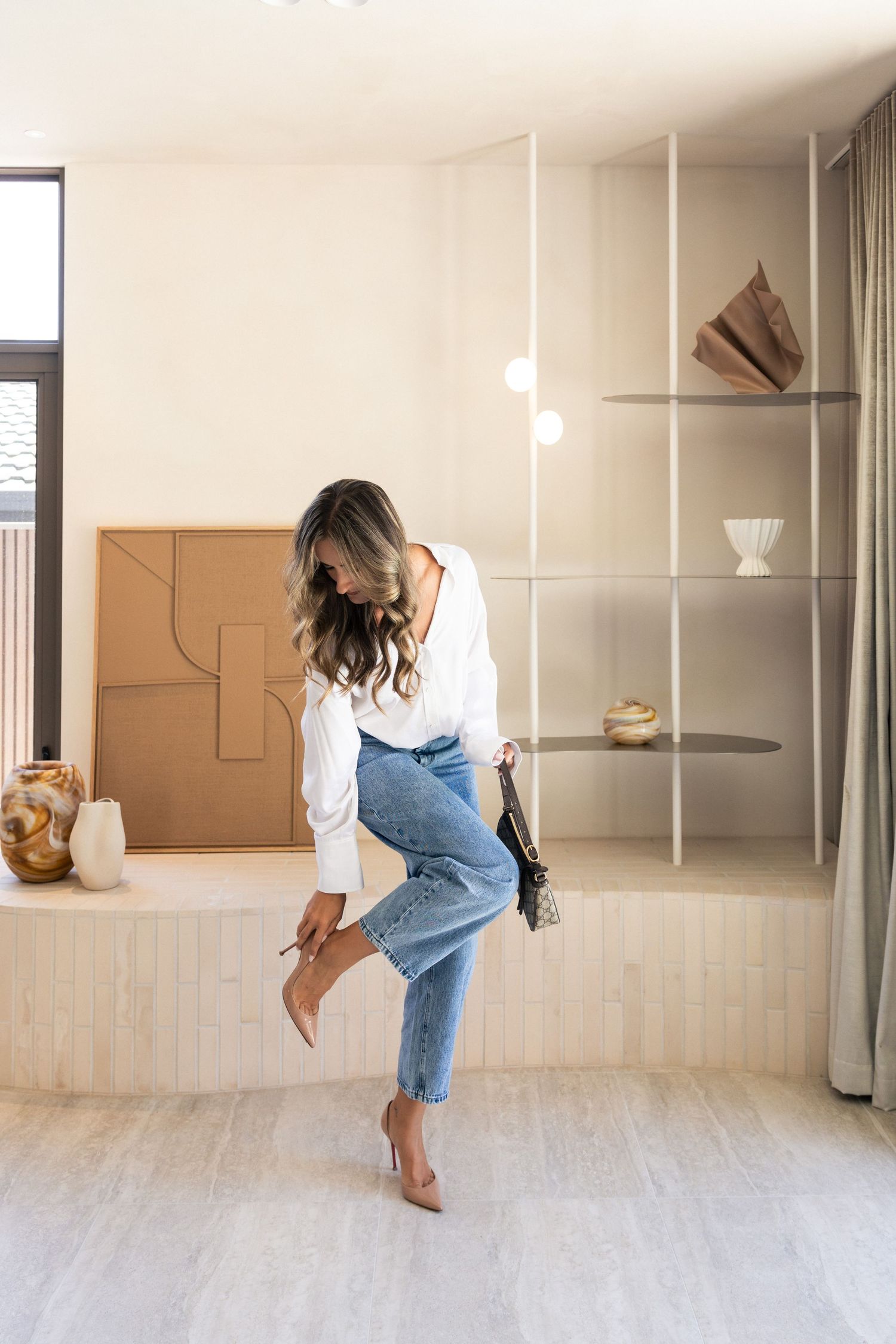 Woman in white blouse and jeans adjusting her heel in a stylish minimalist interior.