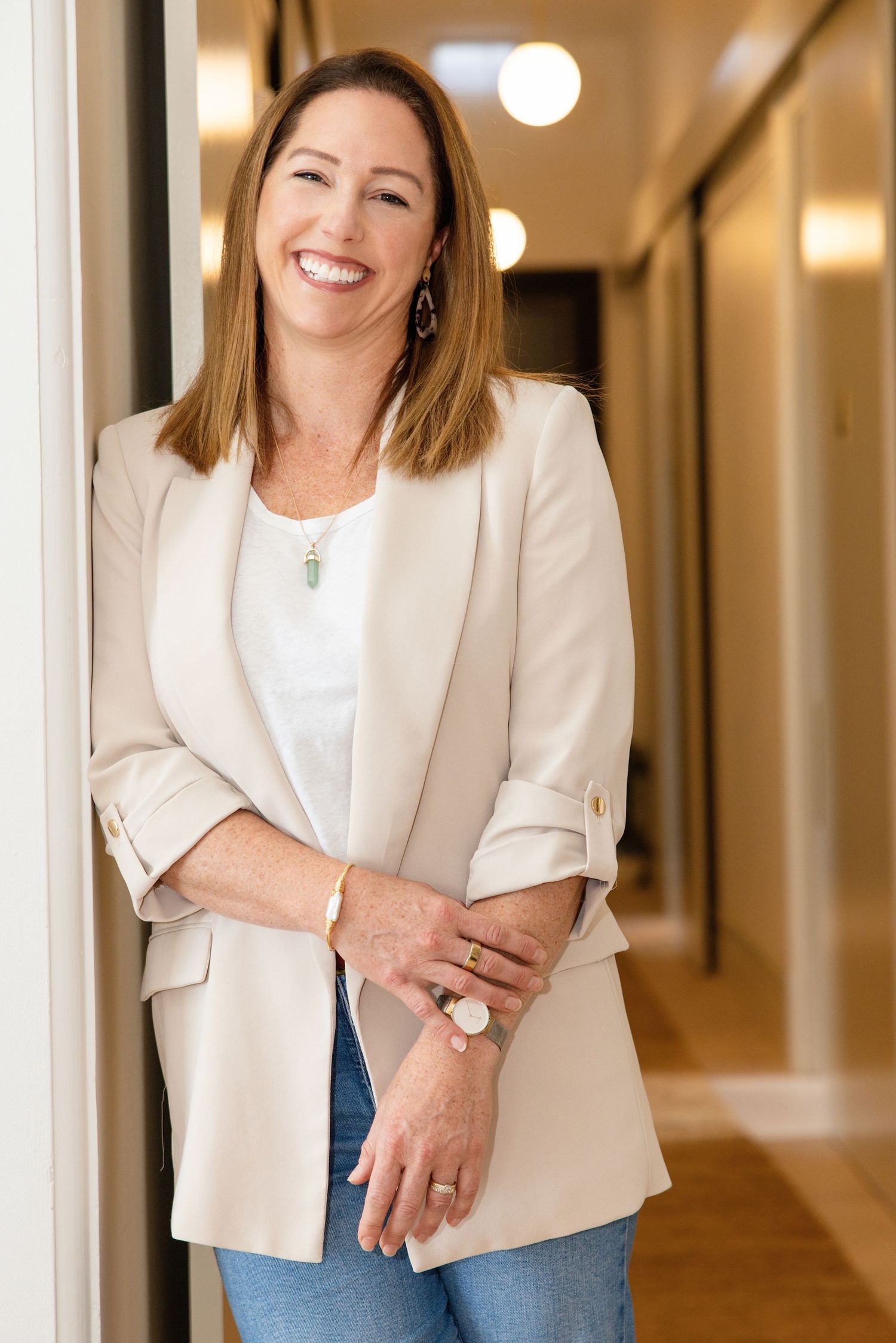 Smiling woman with bob haircut in beige blazer leaning against wall in modern office hallway.