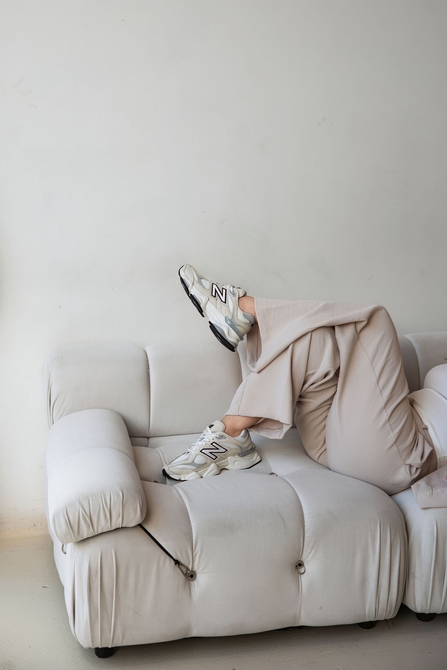 Person lounging on white sofa wearing beige tracksuit and New Balance sneakers with legs raised.