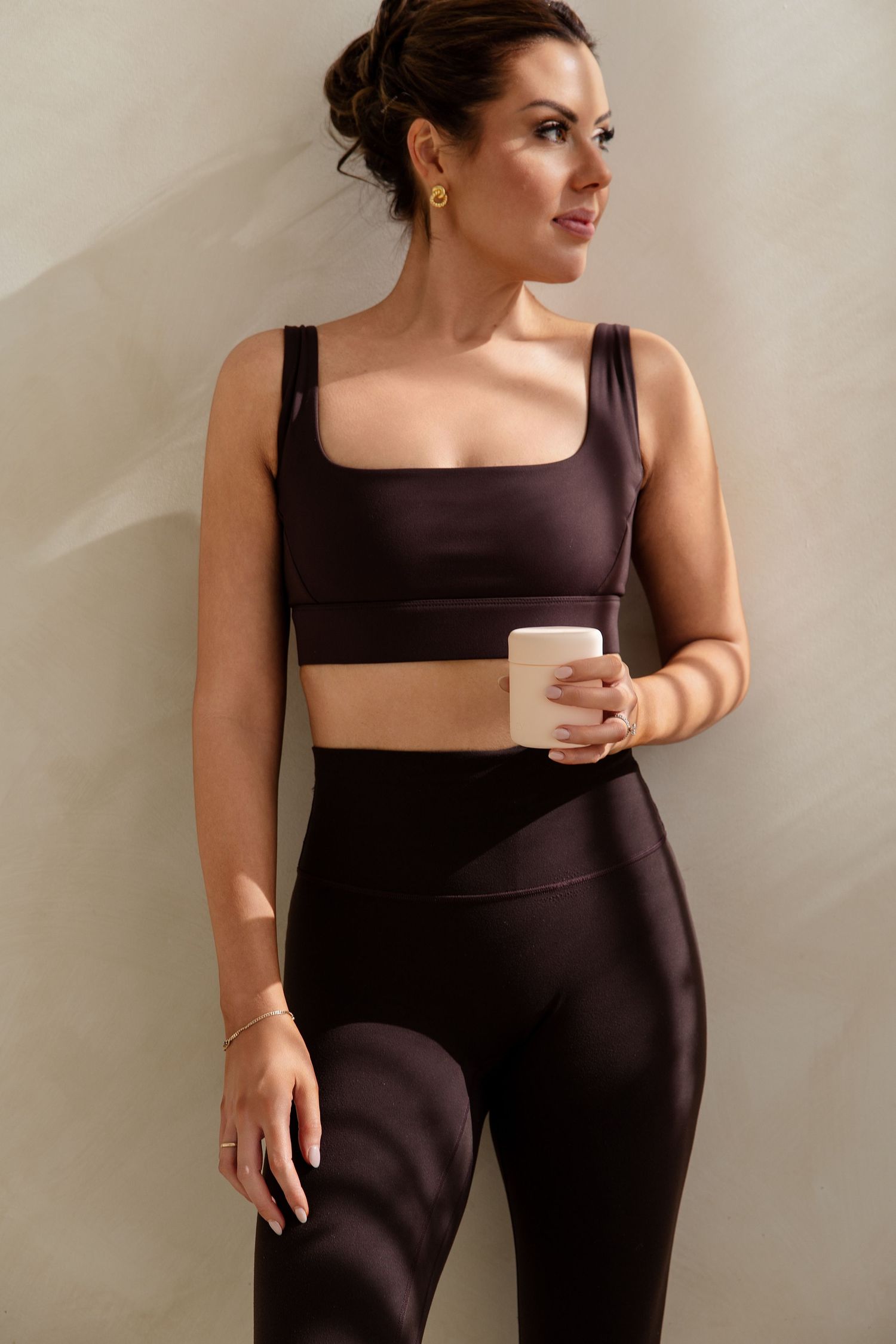 Woman in dark brown satin crop top and high-waist leggings holding a white mug.