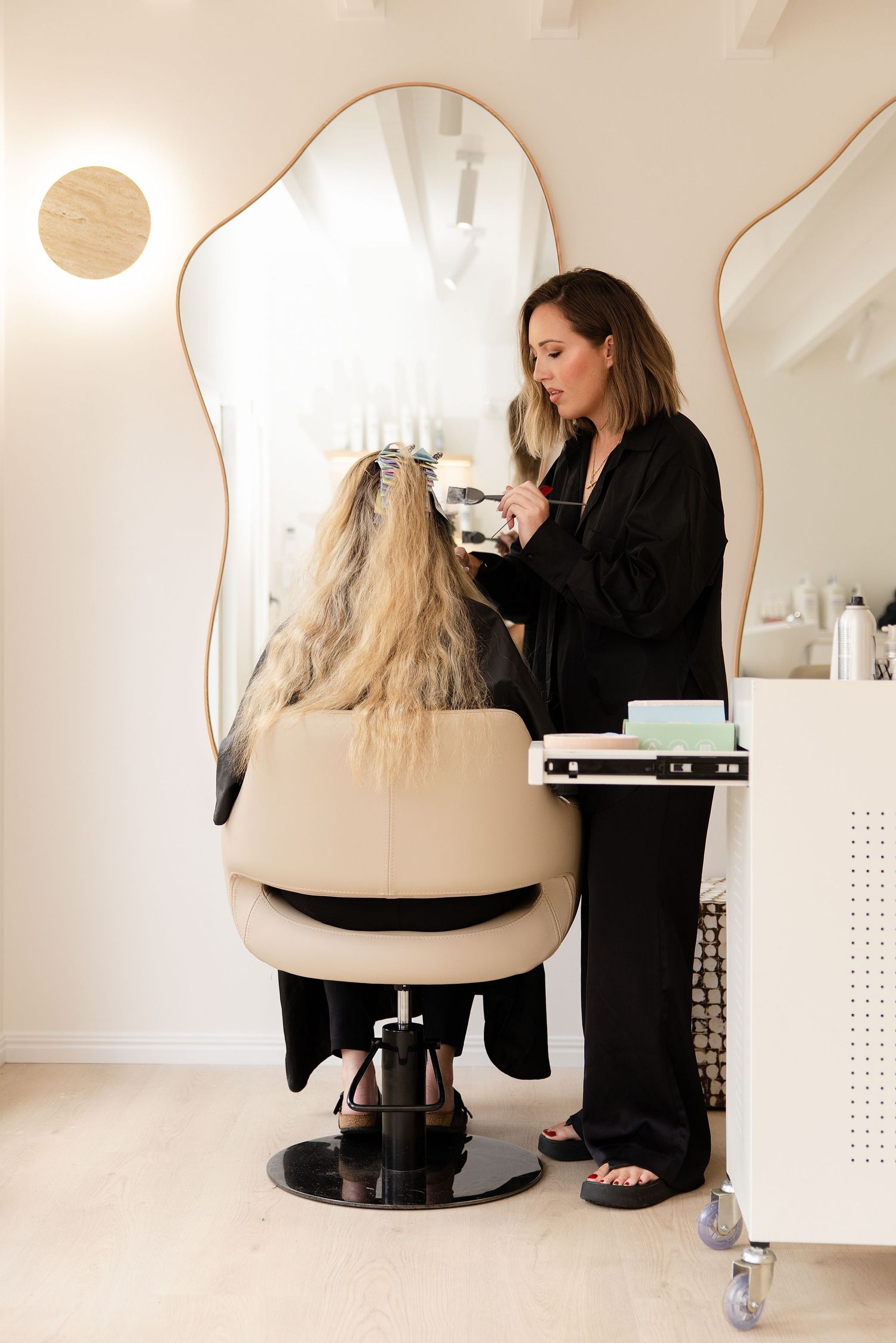 Hair stylist in black robe styling blonde client's hair in modern salon with wavy mirror.