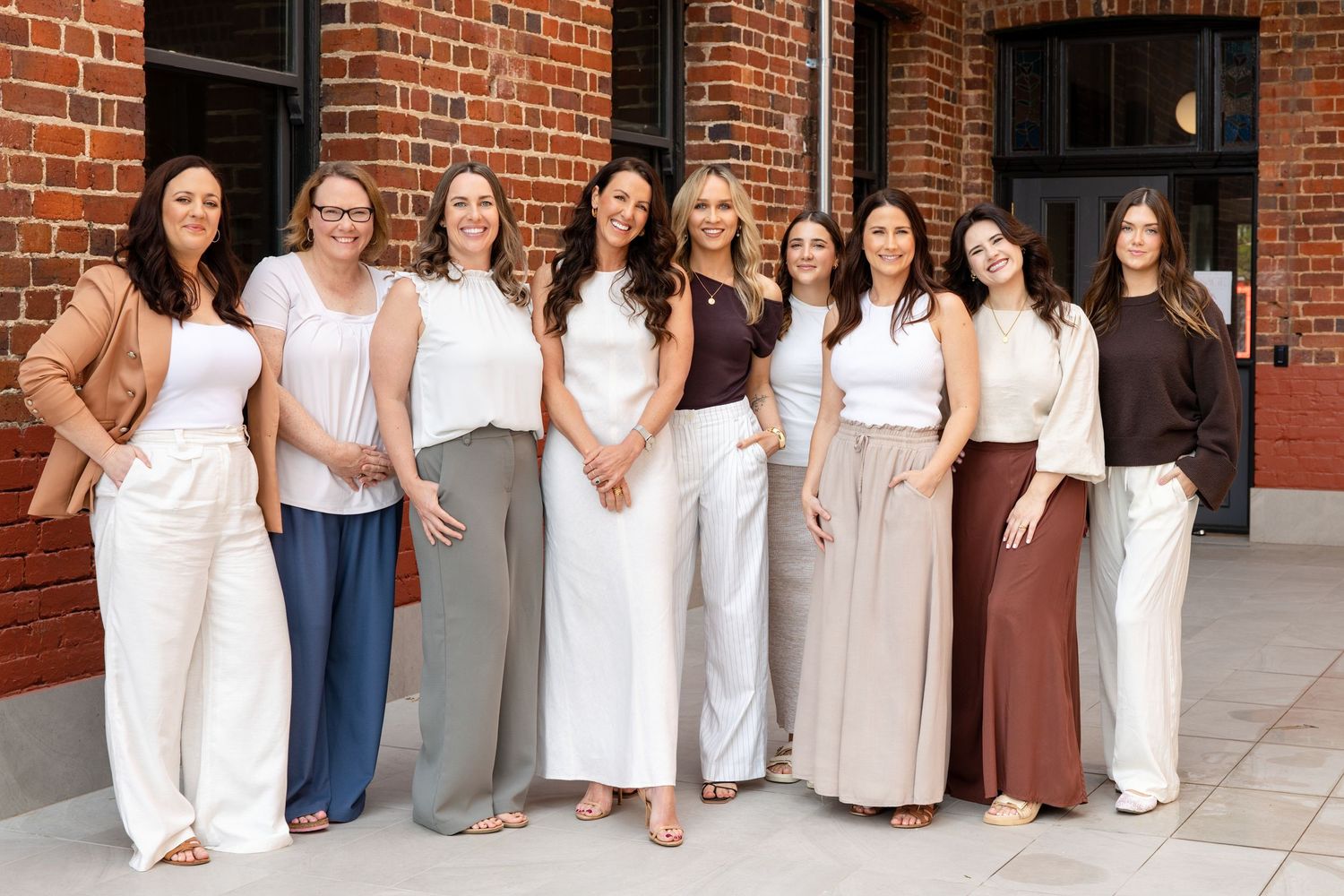 Nine women in neutral outfits smiling together outdoors in front of a brick building.