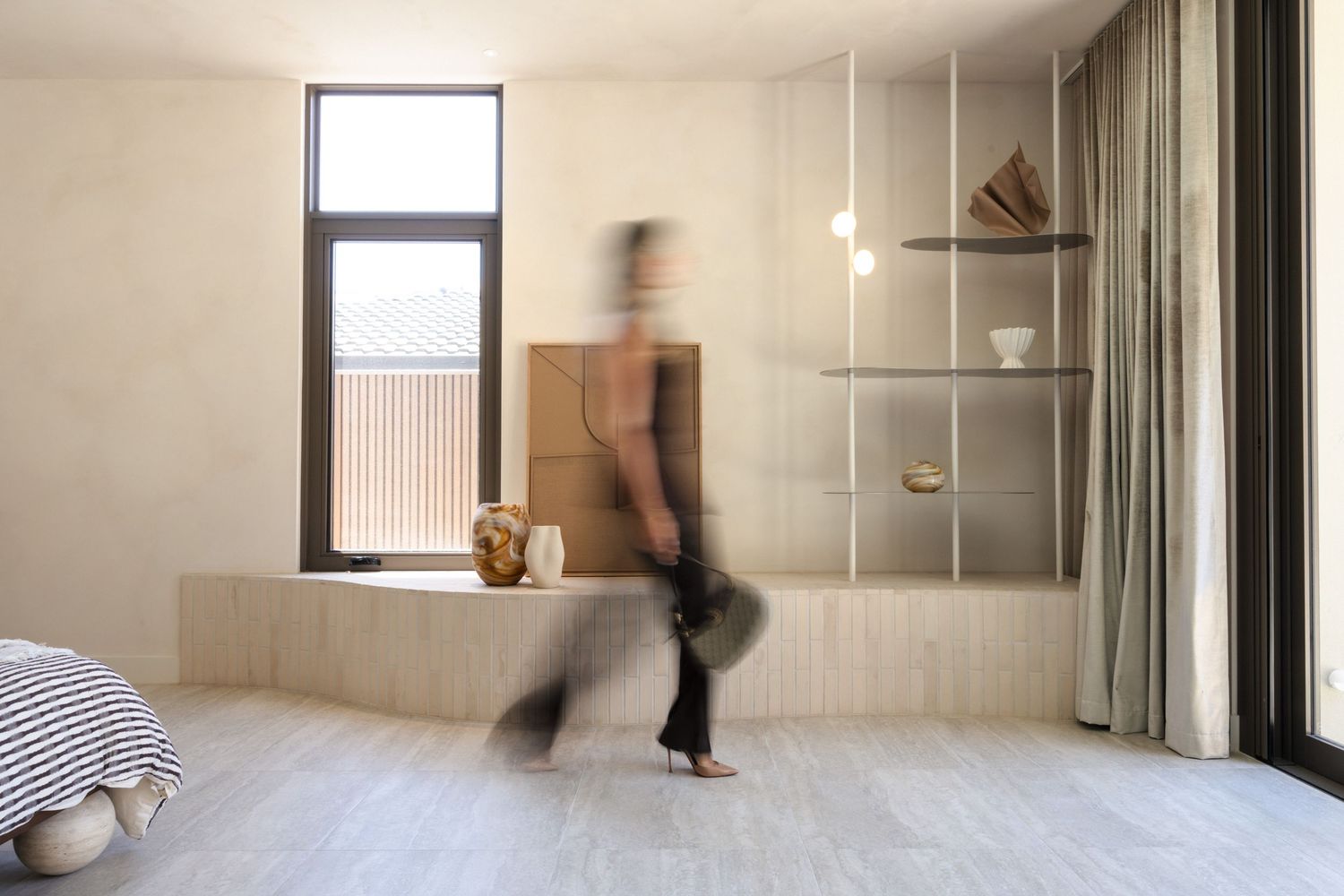 Blurred figure walking through a minimalist room with wooden built-ins, open shelving, and soft neutral tones.