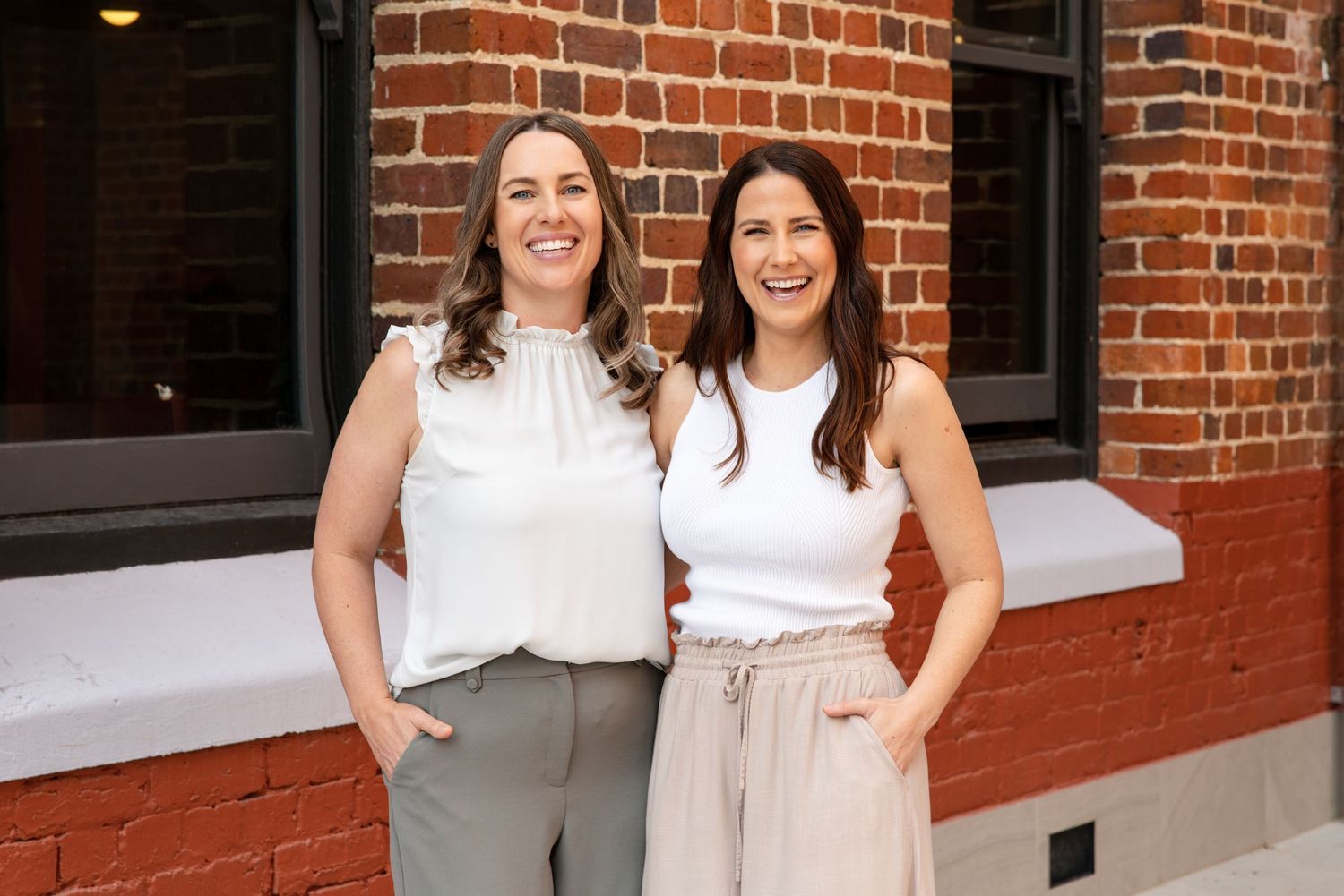Two smiling women in casual white tops pose together outdoors against a red brick wall.