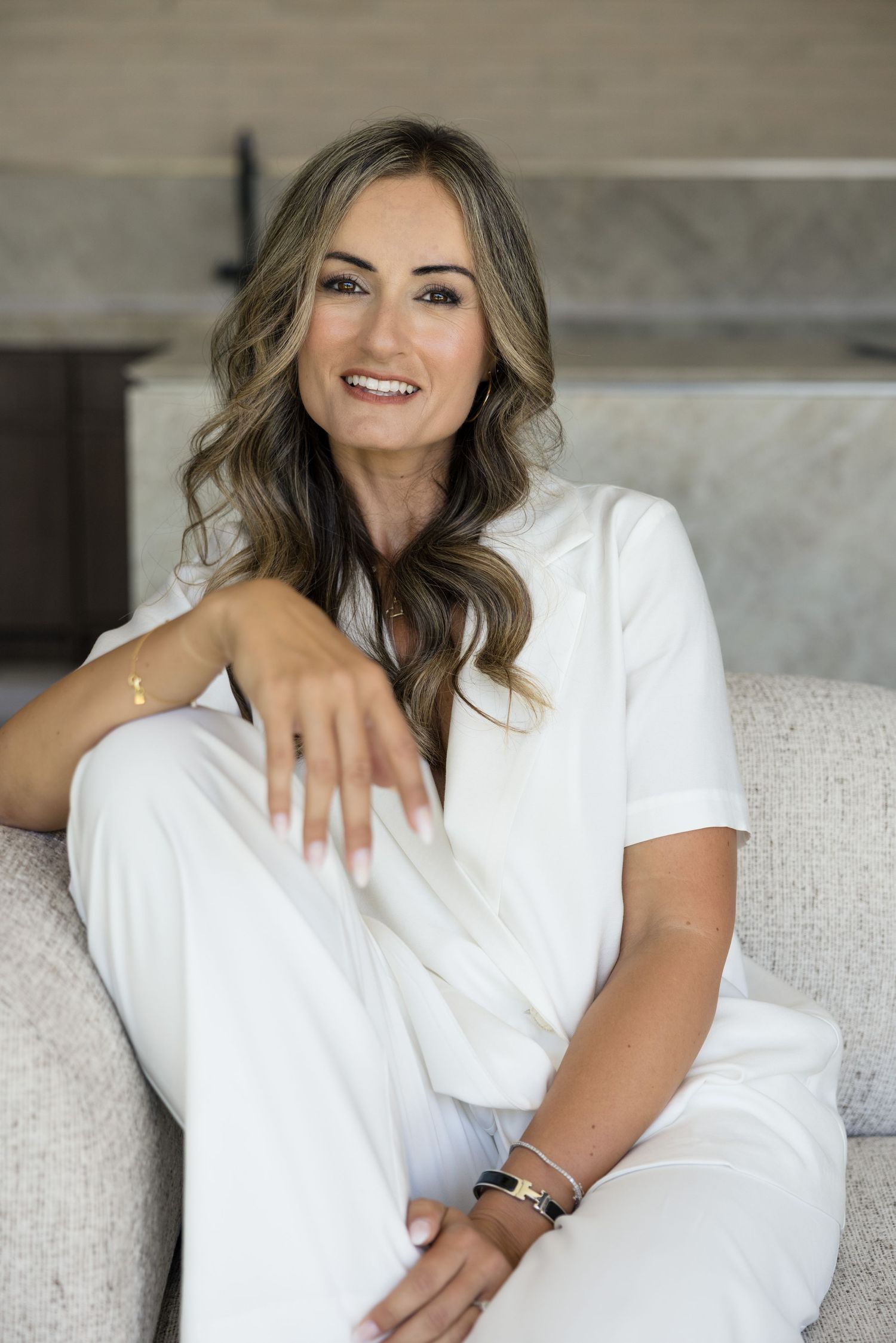 Smiling woman in white outfit sitting relaxed on a neutral sofa in a modern interior.
