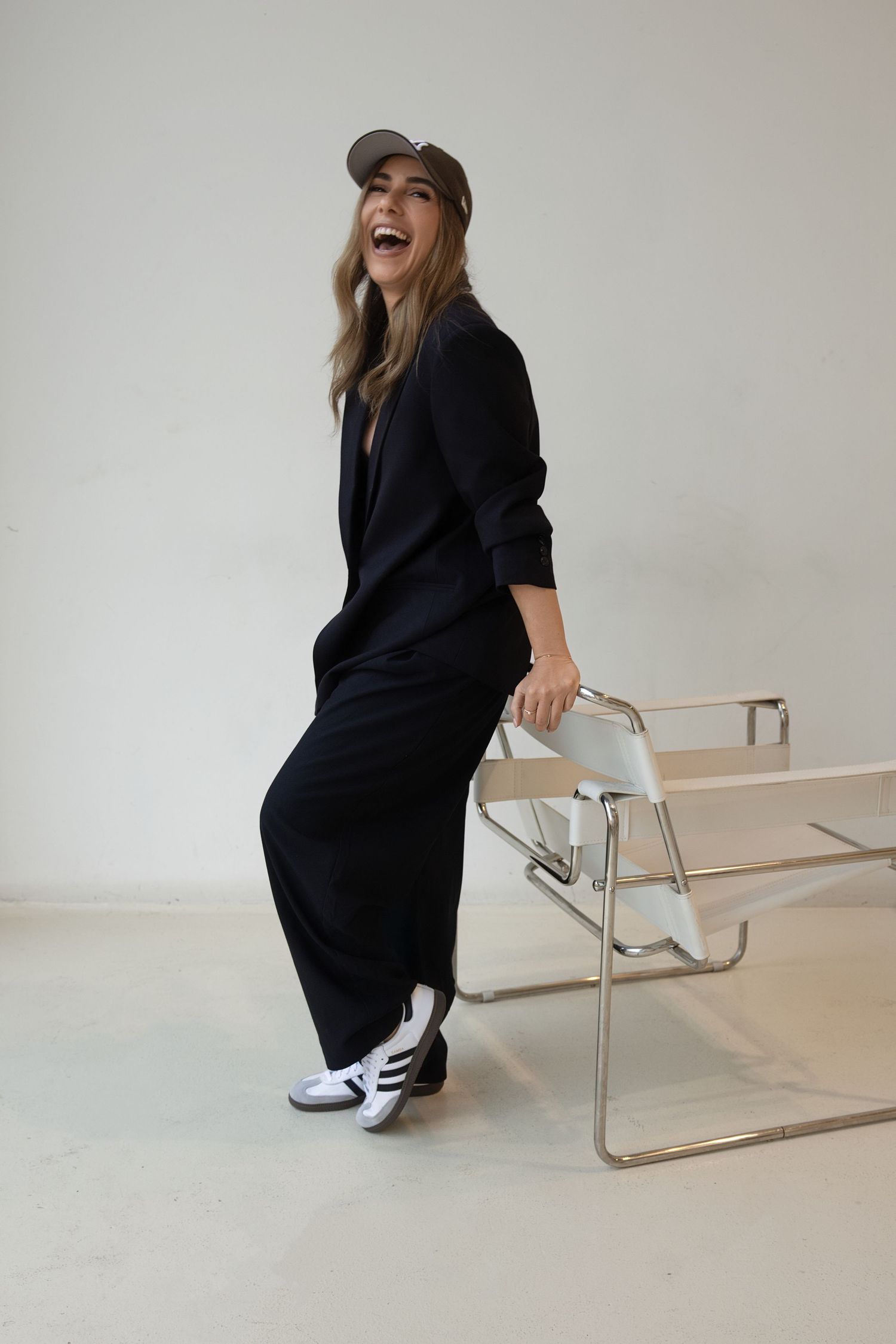 Woman in black jumpsuit and cap laughing while leaning on a modern chrome chair against white background.