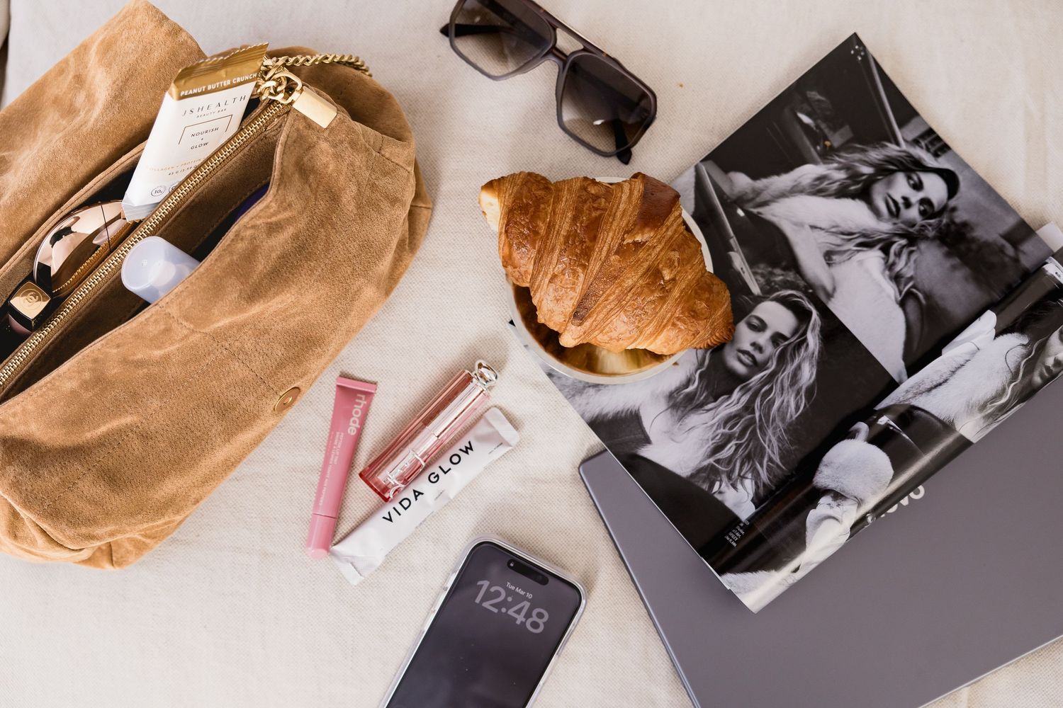 Flatlay of a croissant, sunglasses, brown bag, phone, and vintage black and white photographs on light surface.
