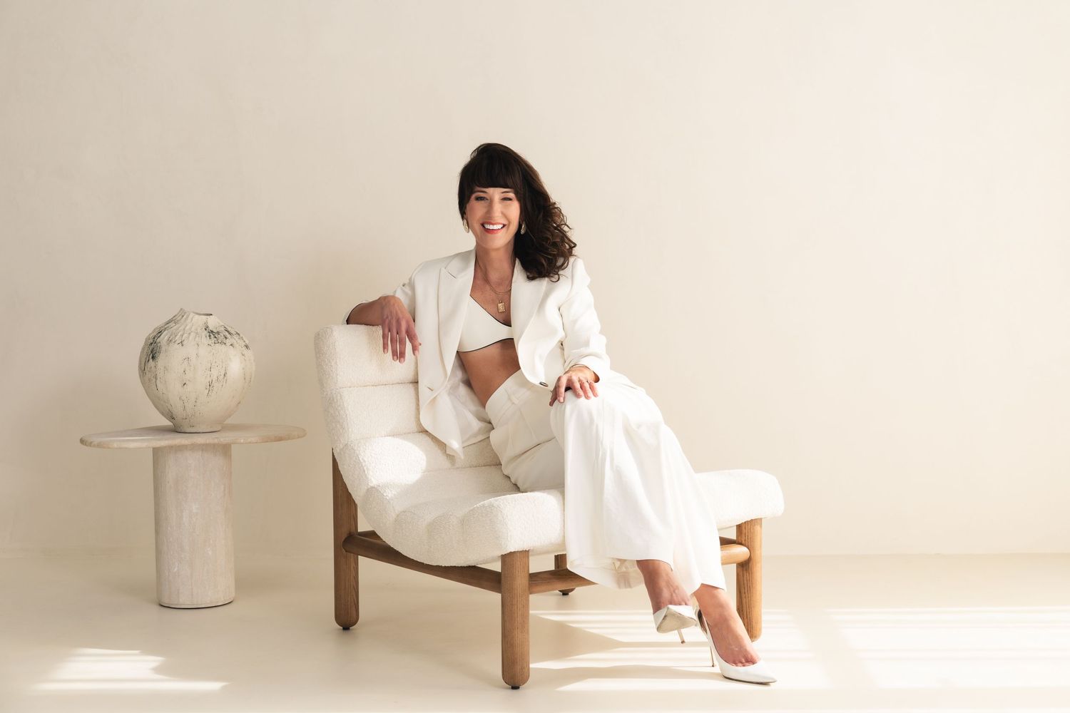 A white outfit is photographed with minimalist decor and wooden furniture in a bright studio setting.