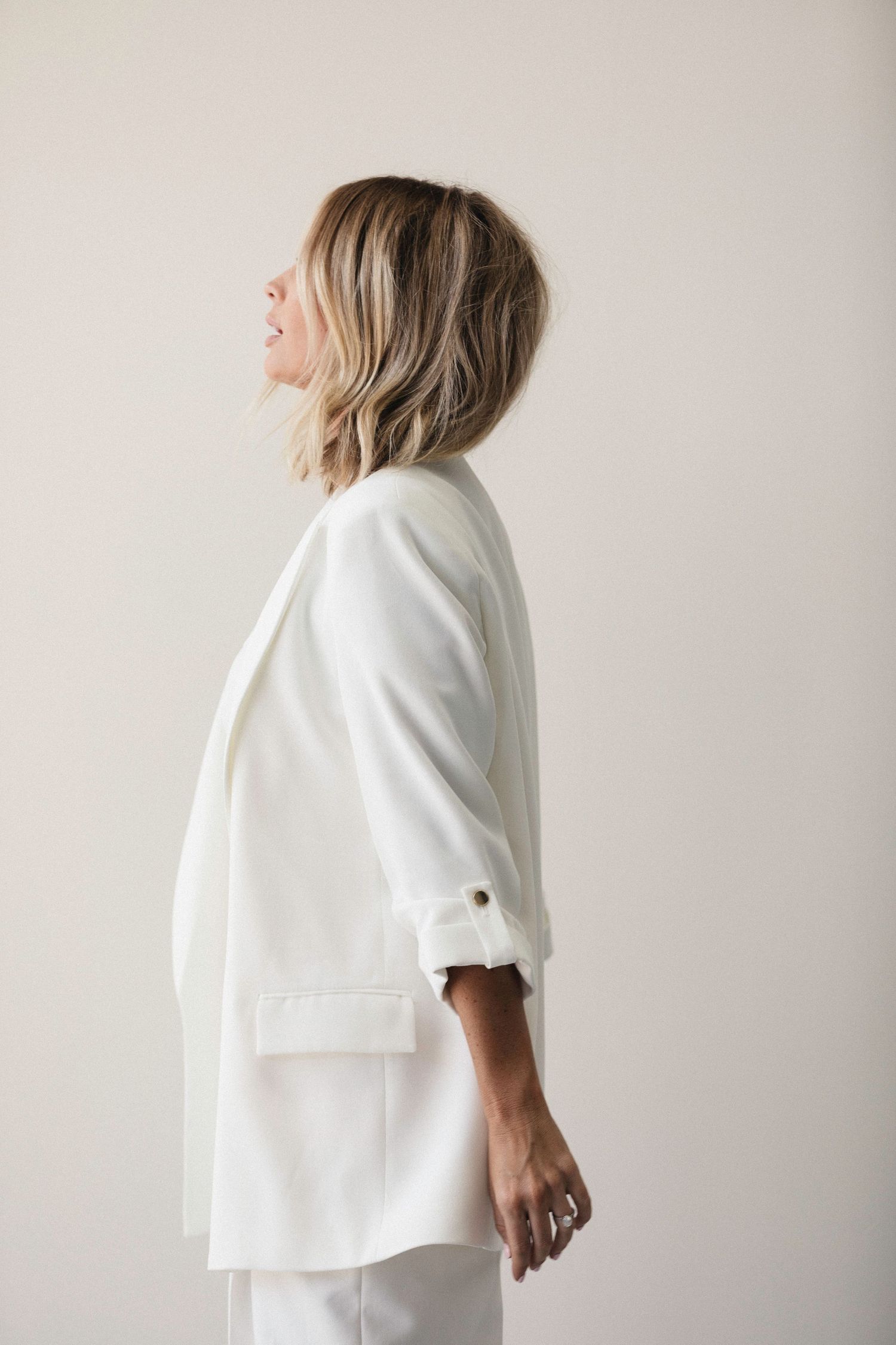Woman in white blazer and pants stands in profile against light background.