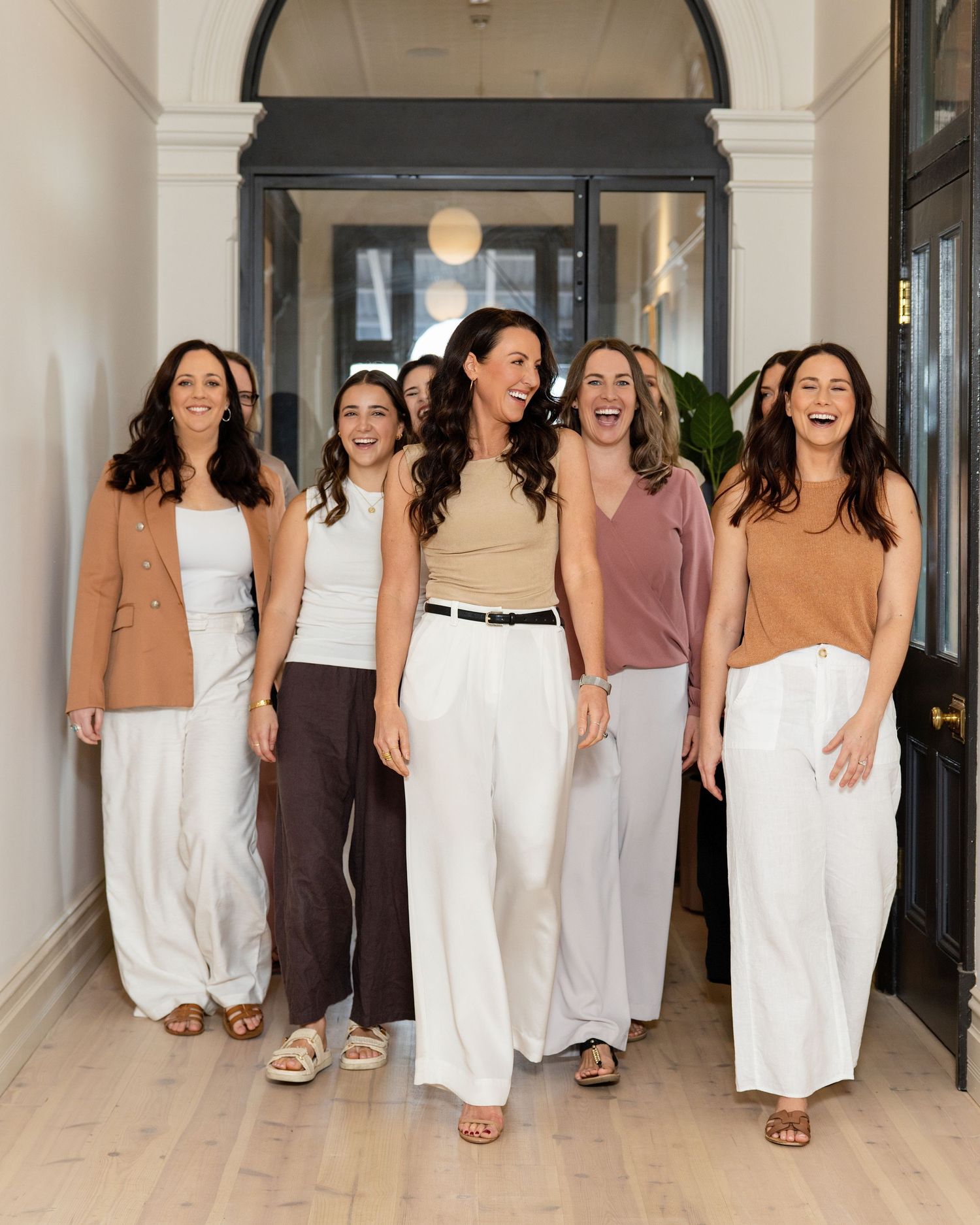 Six smiling women in neutral tones walking confidently down a bright modern hallway together.