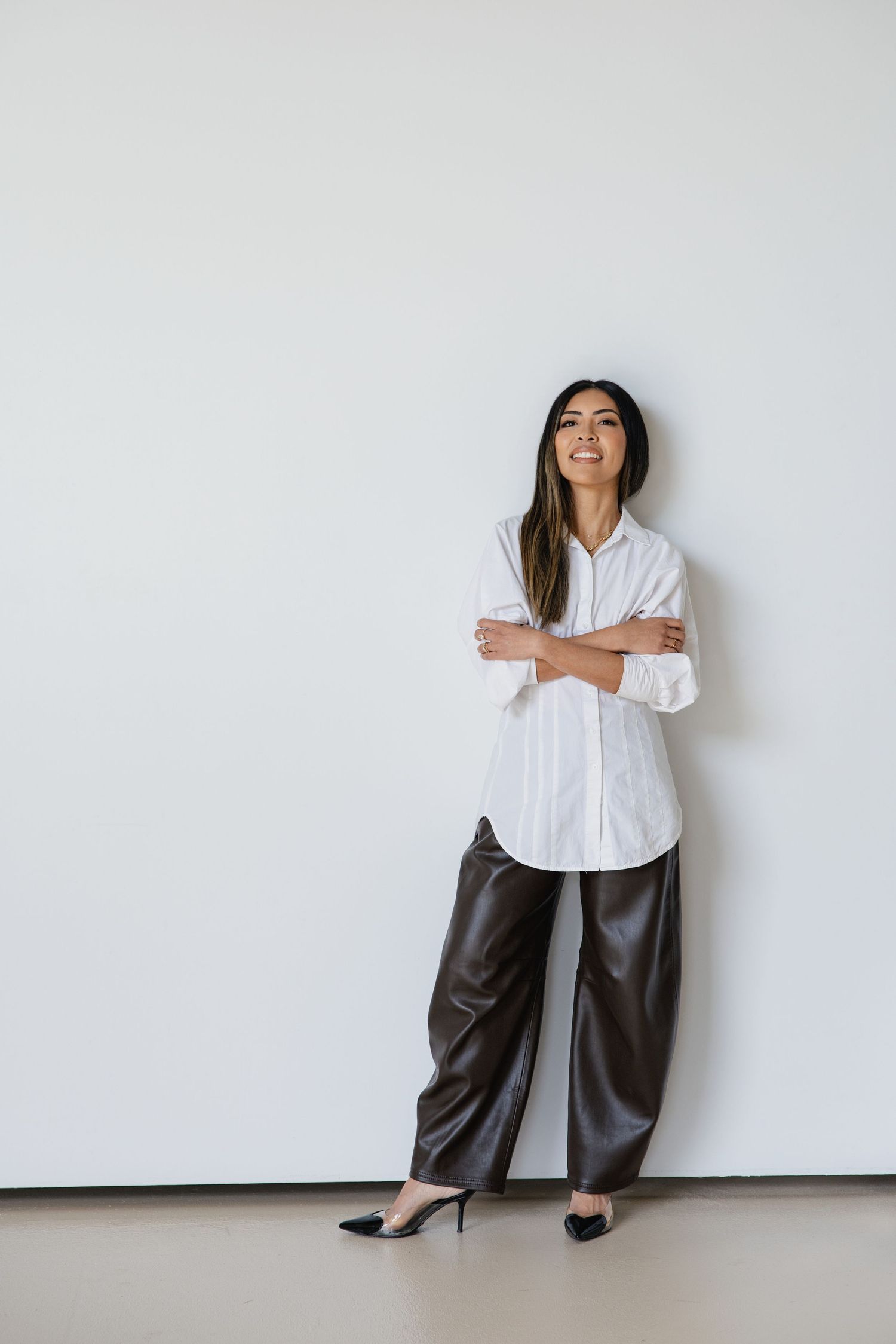 Woman in white button-up shirt and dark leather wide-leg pants with black heels, standing confidently against a white wall.