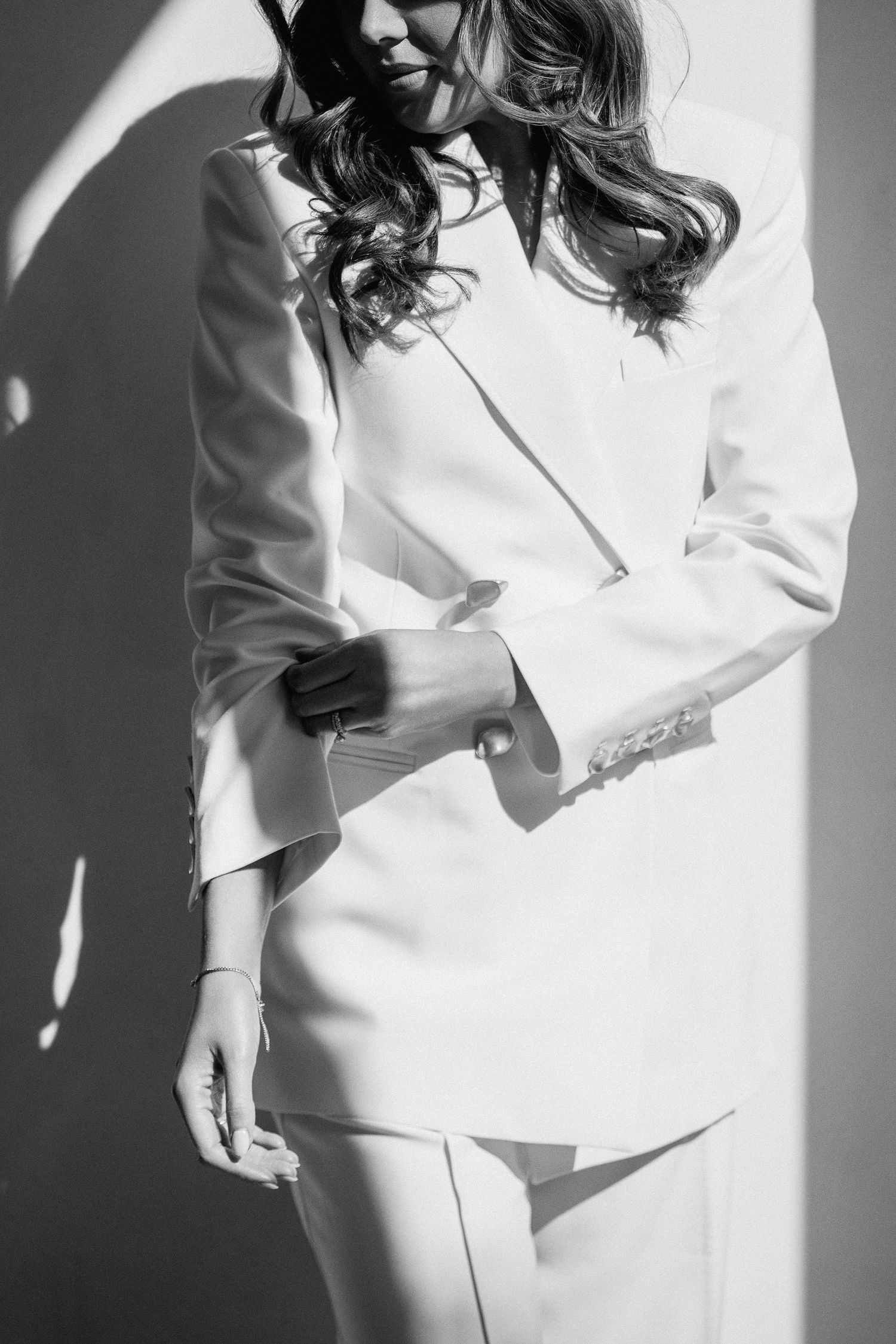 Black and white photo of a woman in a white blazer suit, posing elegantly against a sunlit wall.