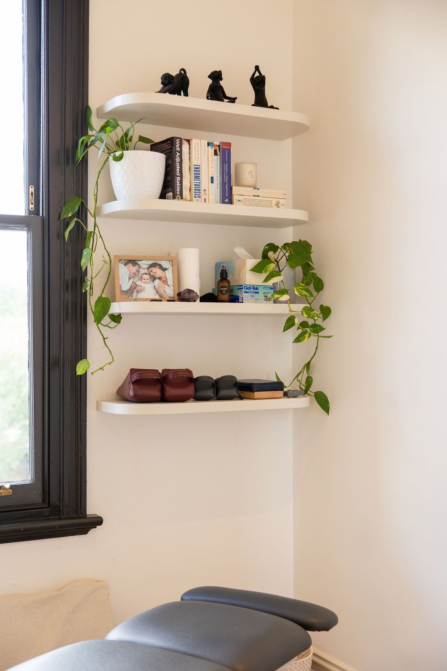 White floating shelves with books, plants, and decor beside a dark-framed window above a treatment table.