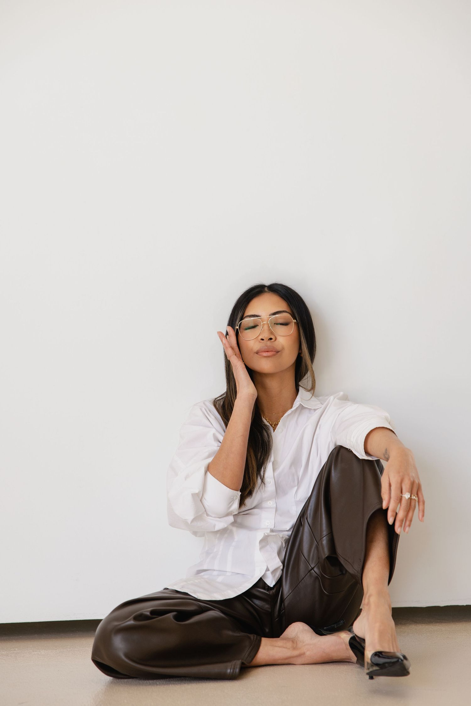 Woman in white shirt and brown leather pants sitting on floor, leaning against white wall, wearing glasses.