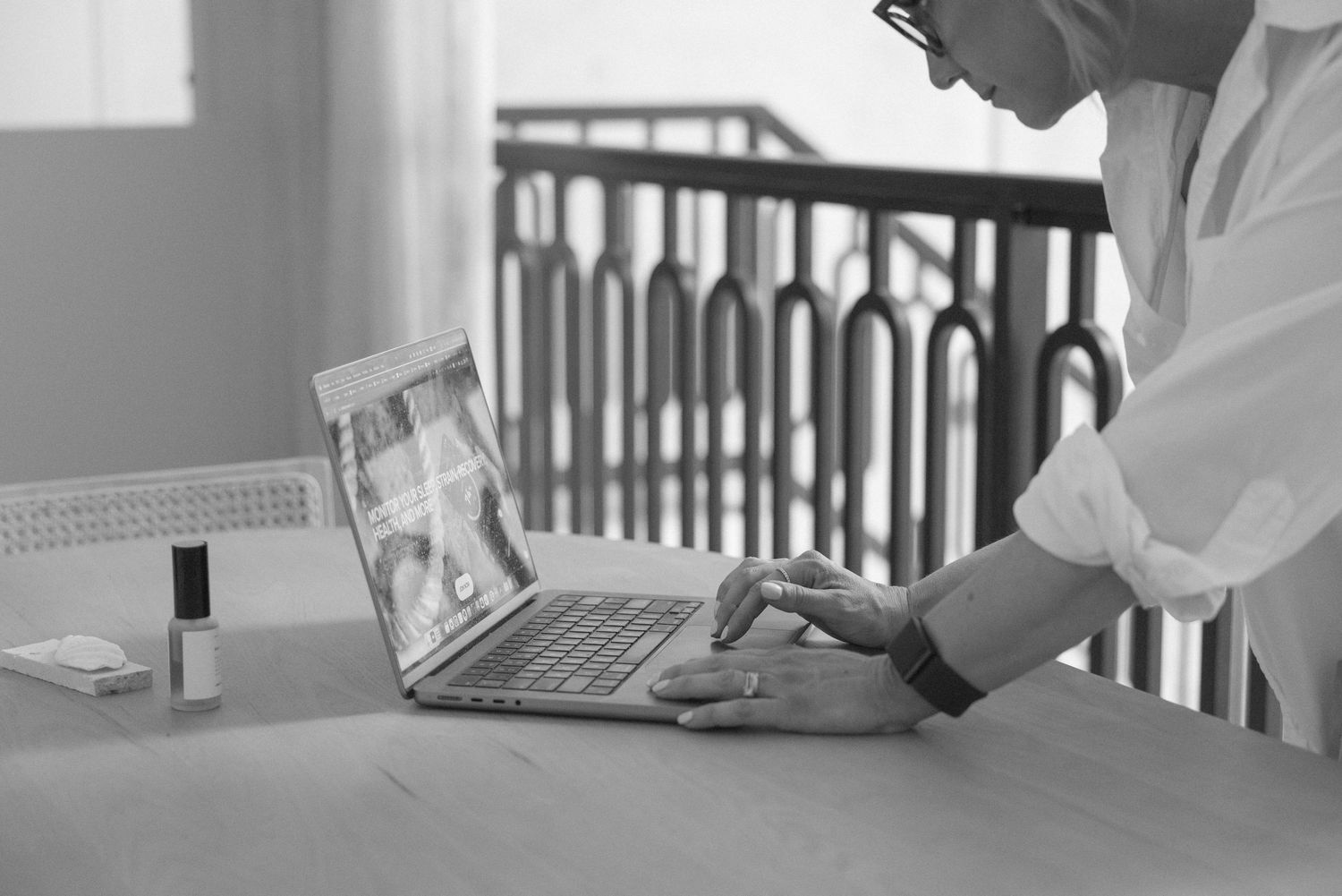 Person in white shirt typing on laptop at desk with nail polish bottles nearby, black and white photo.