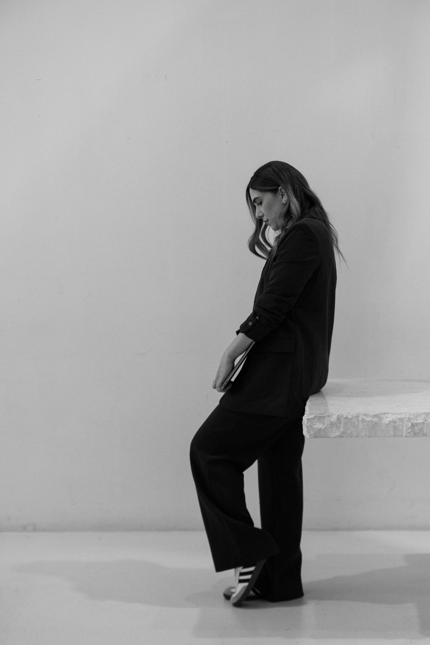 Black and white photo of a woman in a dark blazer and wide-leg trousers leaning against a wall.