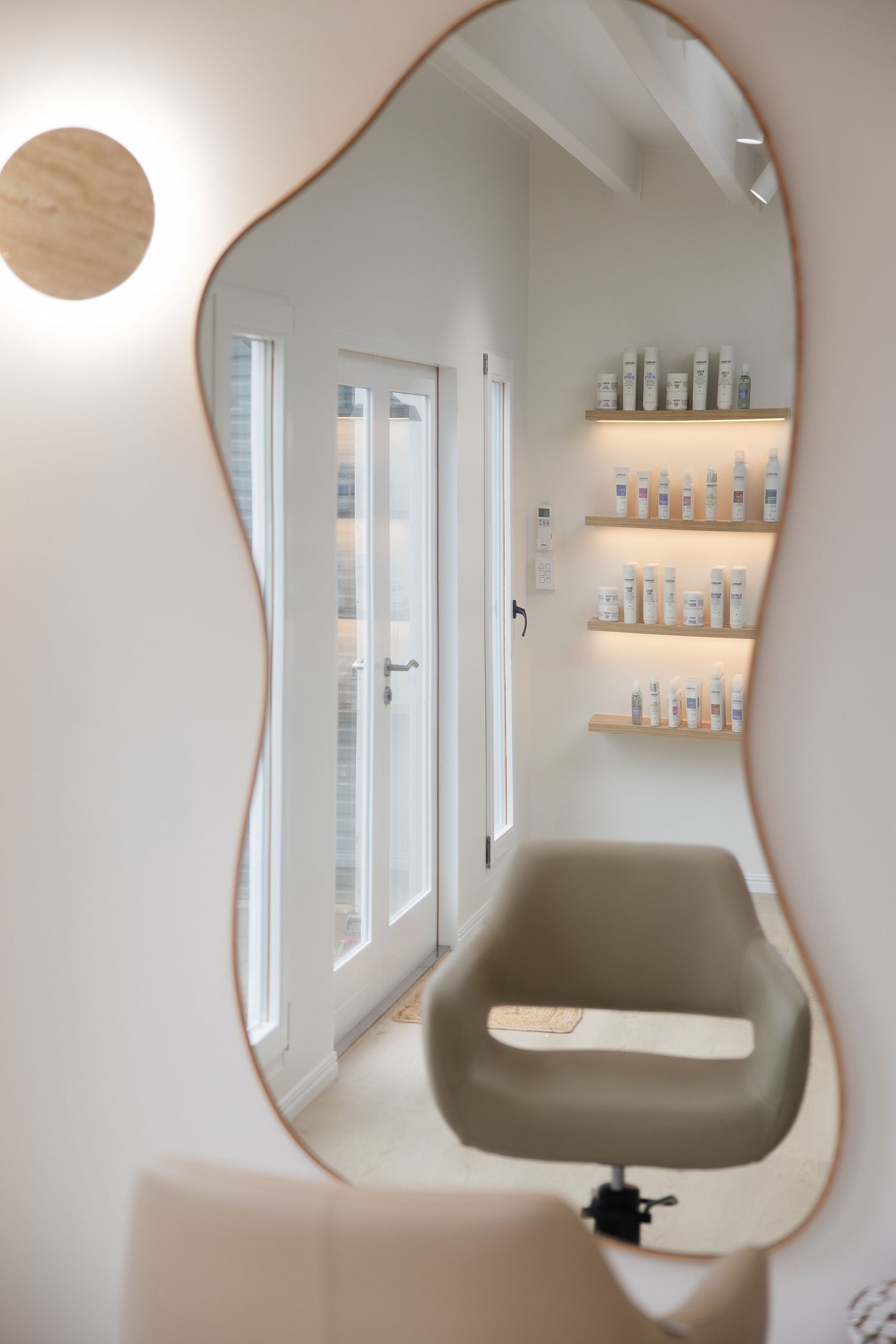 Modern hair salon station with wavy rose gold mirror, olive green styling chair, and illuminated product shelves.