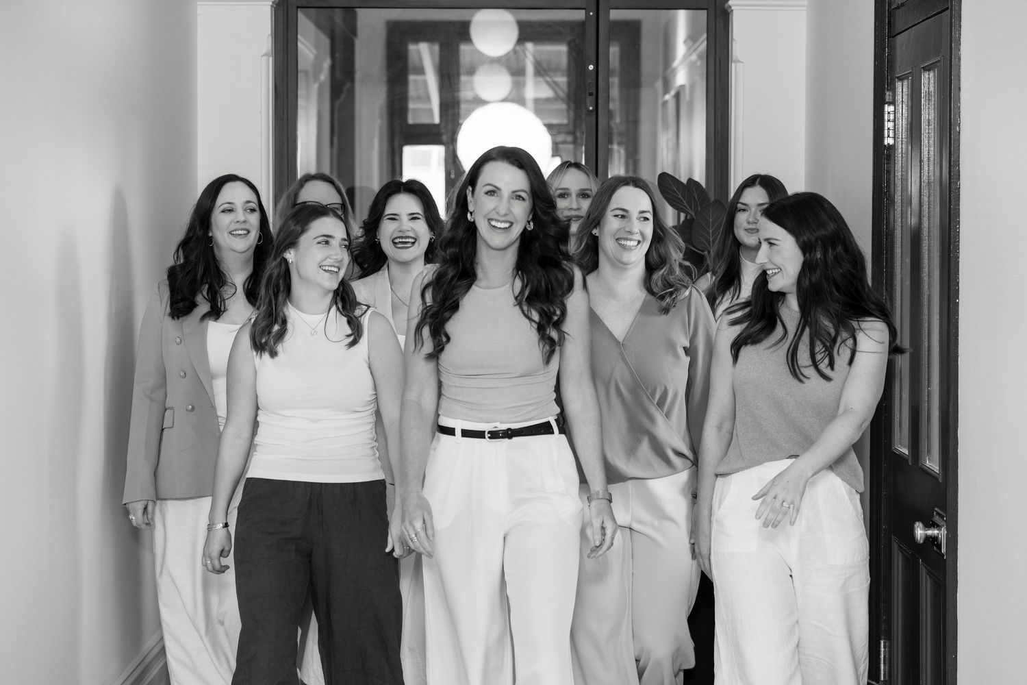 Black and white photo of seven smiling women walking together down a hallway in elegant casual attire.