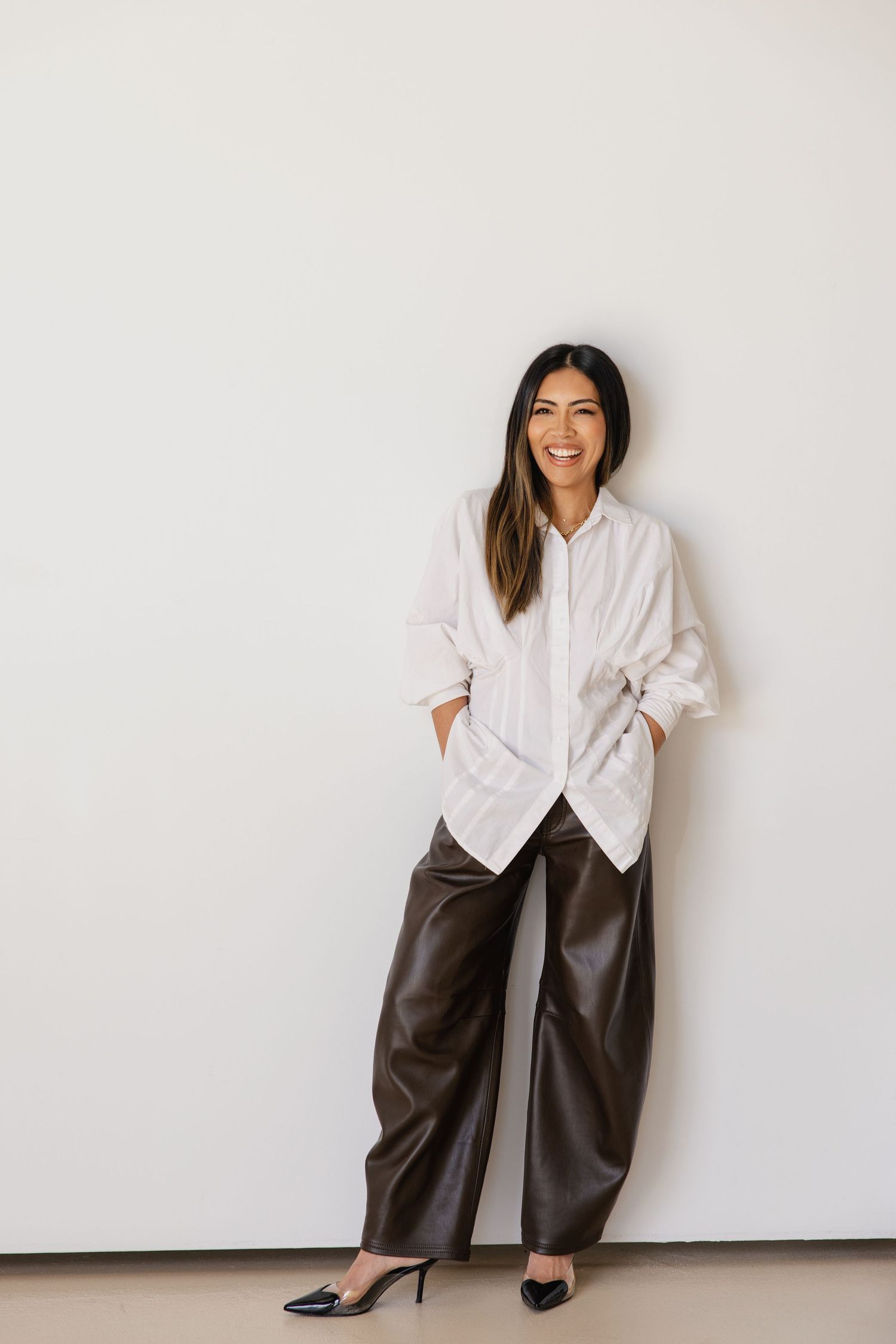 Woman smiling in white button-up shirt and brown leather wide-leg pants with black heels against white wall.