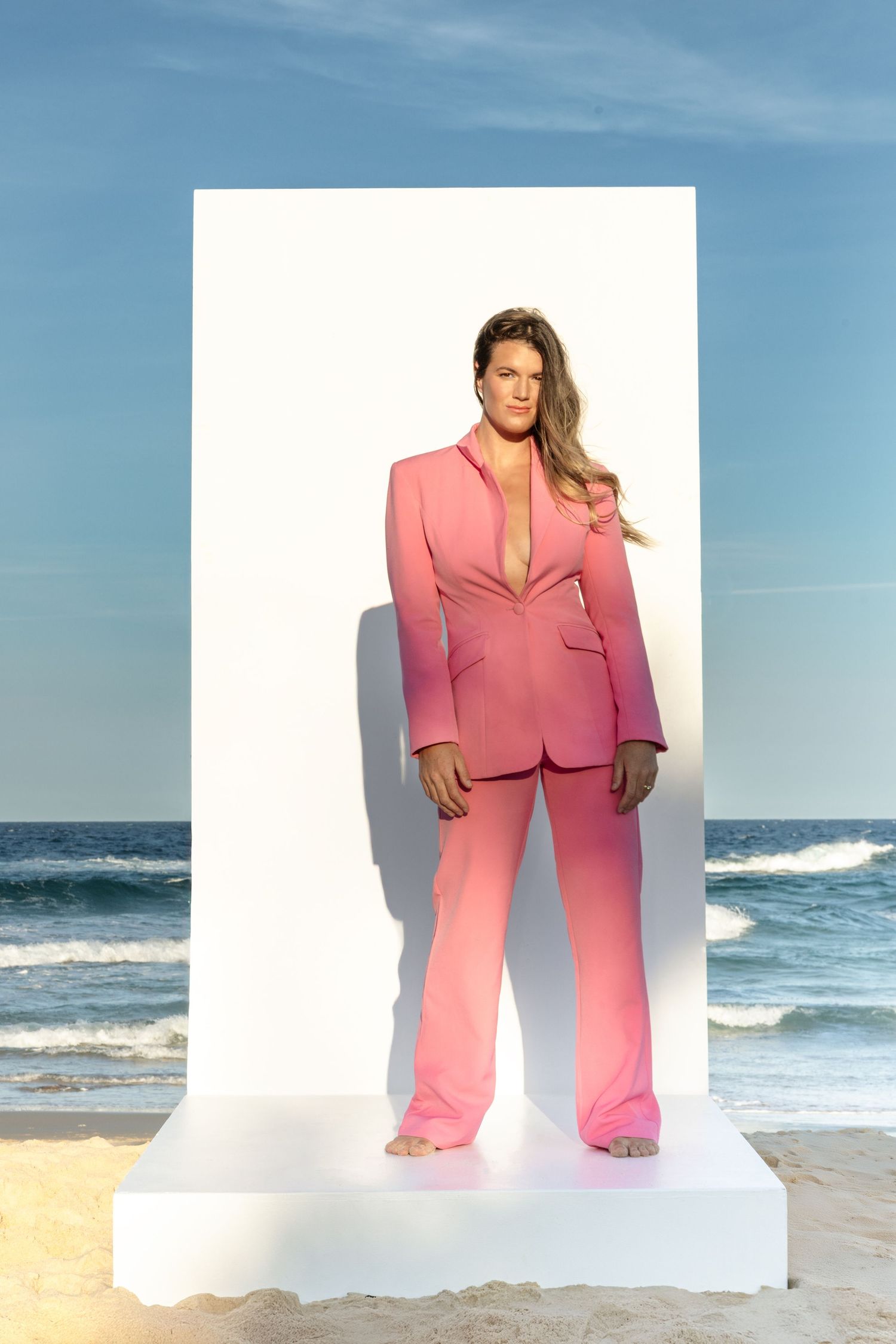 A pink power suit ensemble photographed on a white platform against an ocean backdrop.