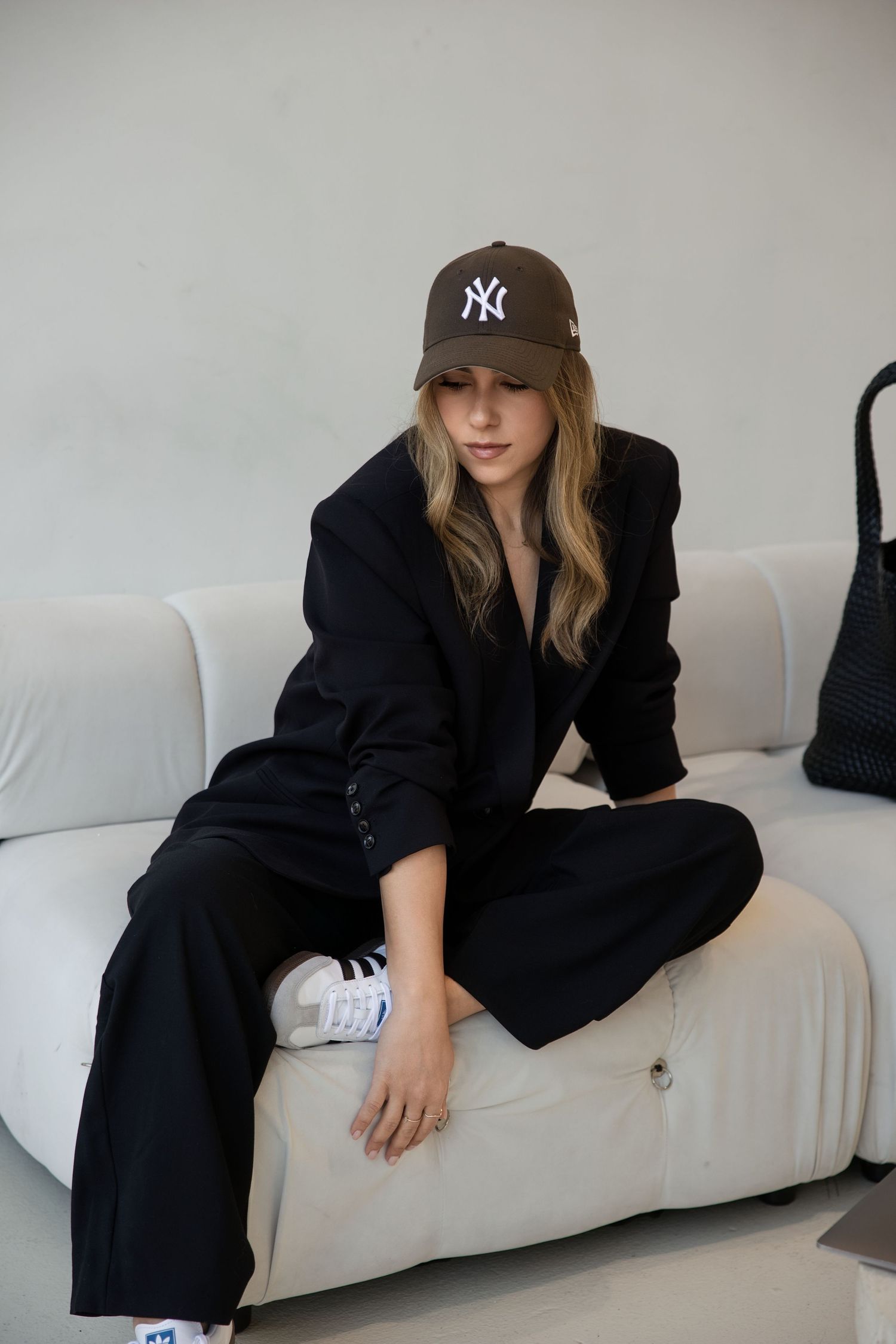 Woman in black blazer and trousers wearing NY Yankees cap sitting on white sofa with Adidas sneakers.