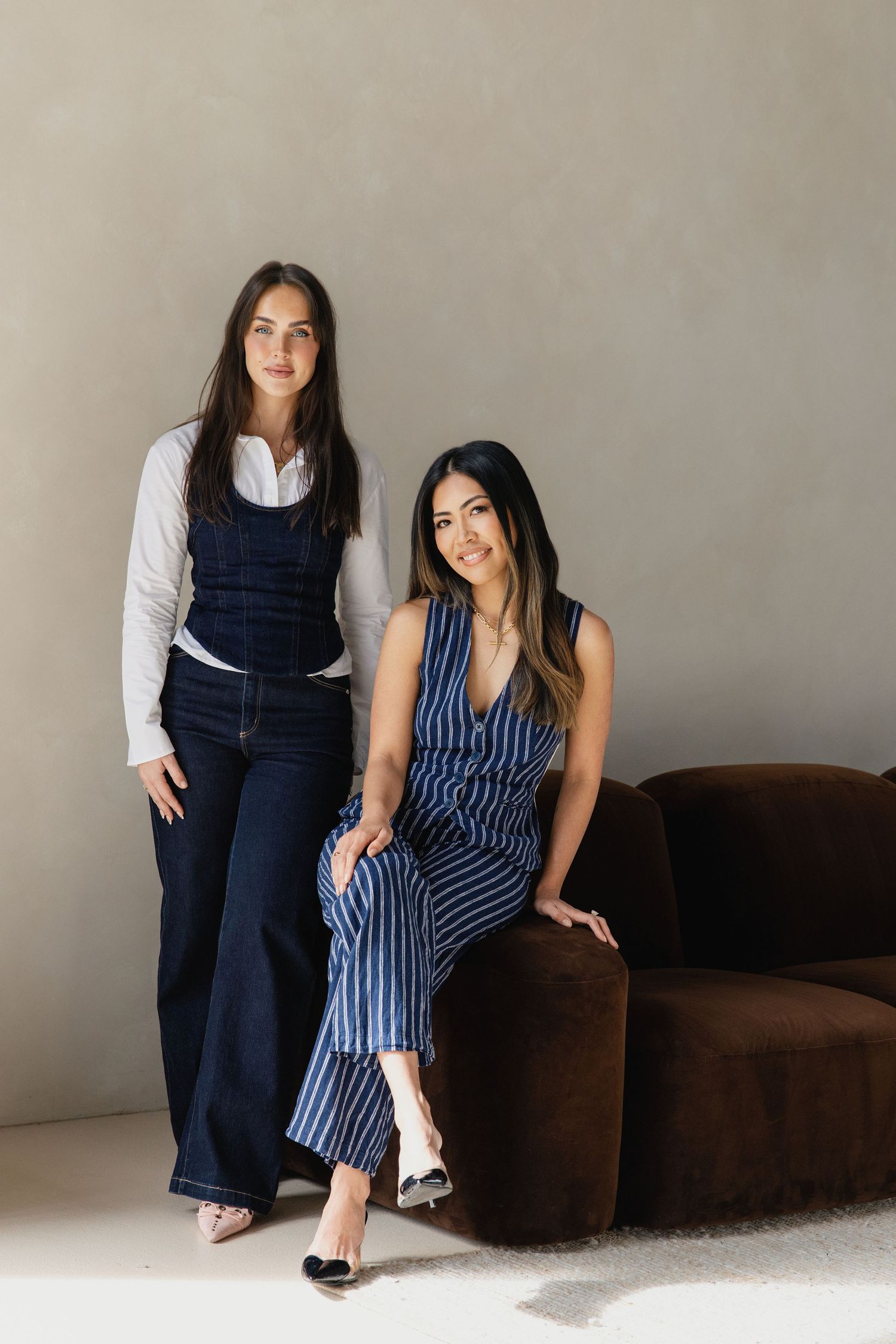 Two stylish women pose in blue outfits, one in denim overalls and one in a striped jumpsuit, against a neutral wall.