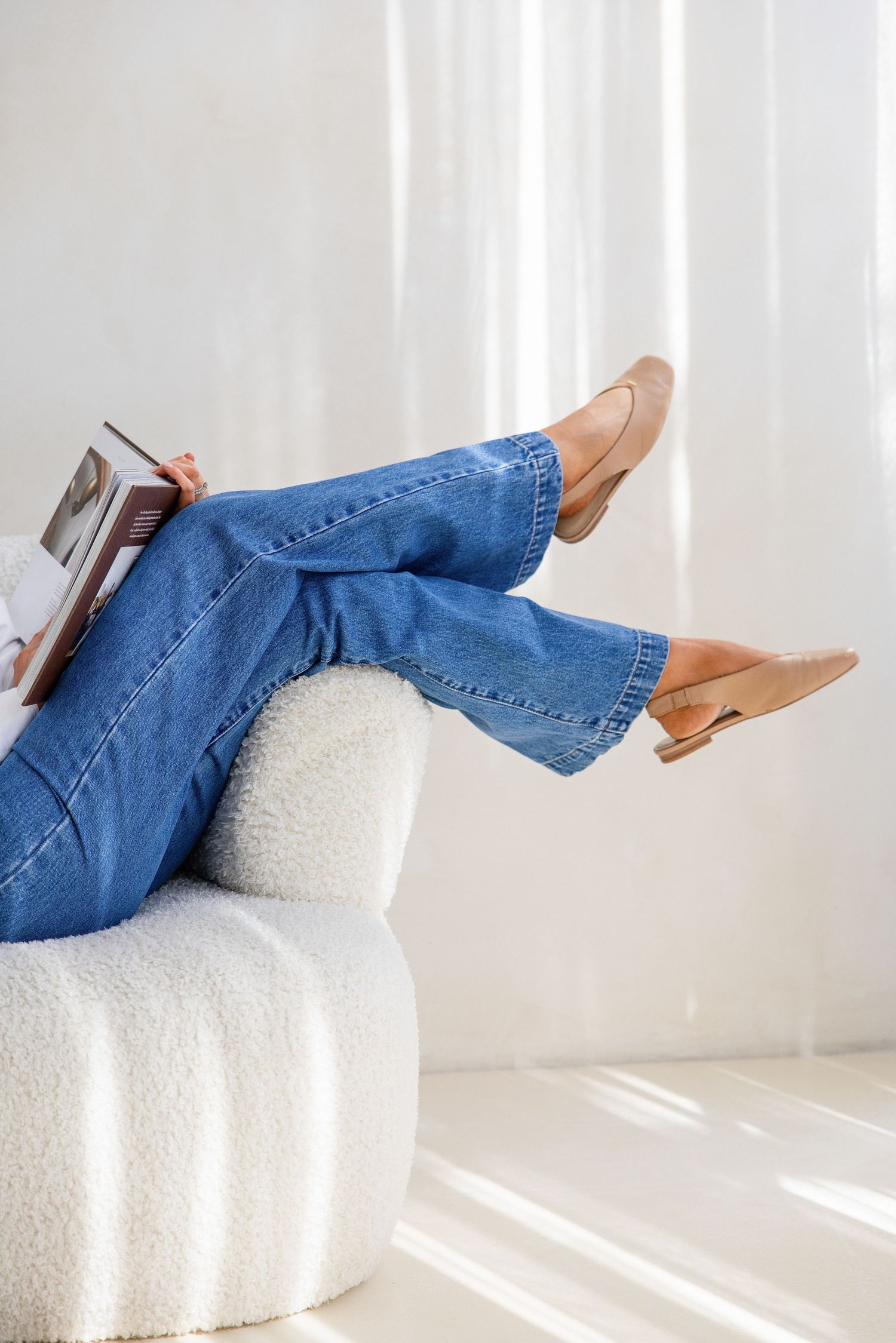 Woman relaxing on a boucle chair with blue wide-leg jeans and tan flats holding a magazine.