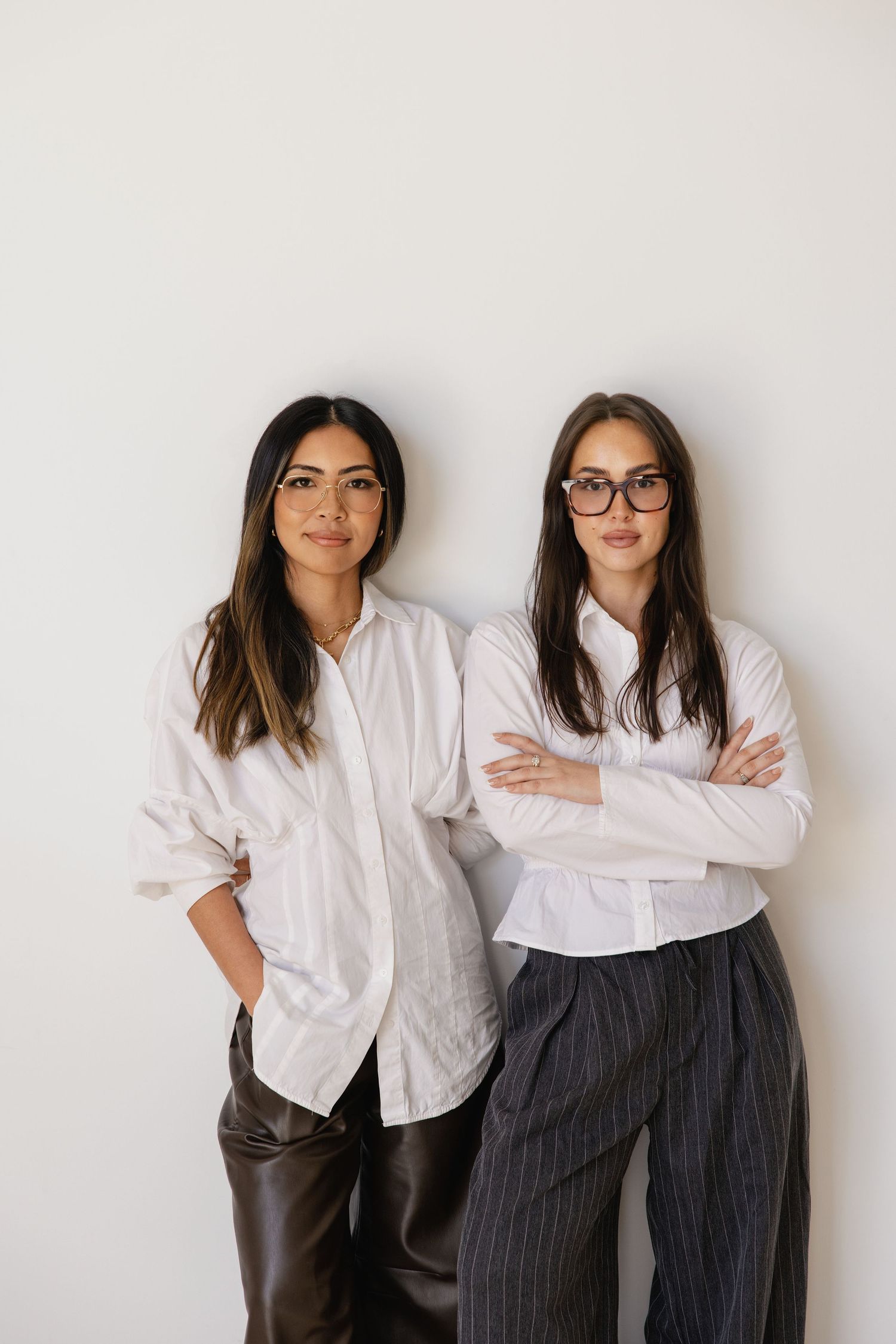 Two professional women in white shirts and glasses posing confidently against a clean white wall.