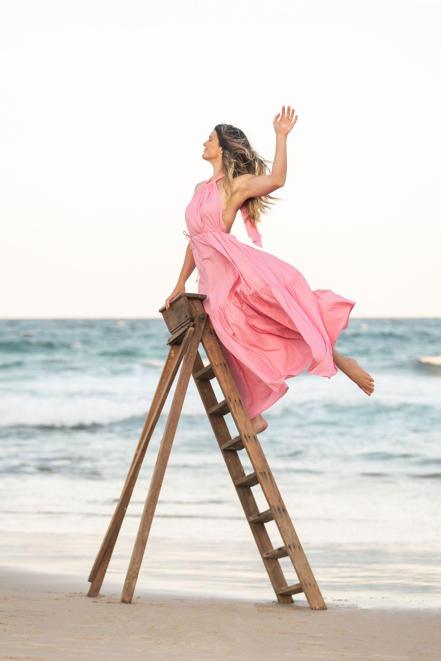 A person in a flowing pink dress poses playfully on a wooden ladder at the beach during sunset.
