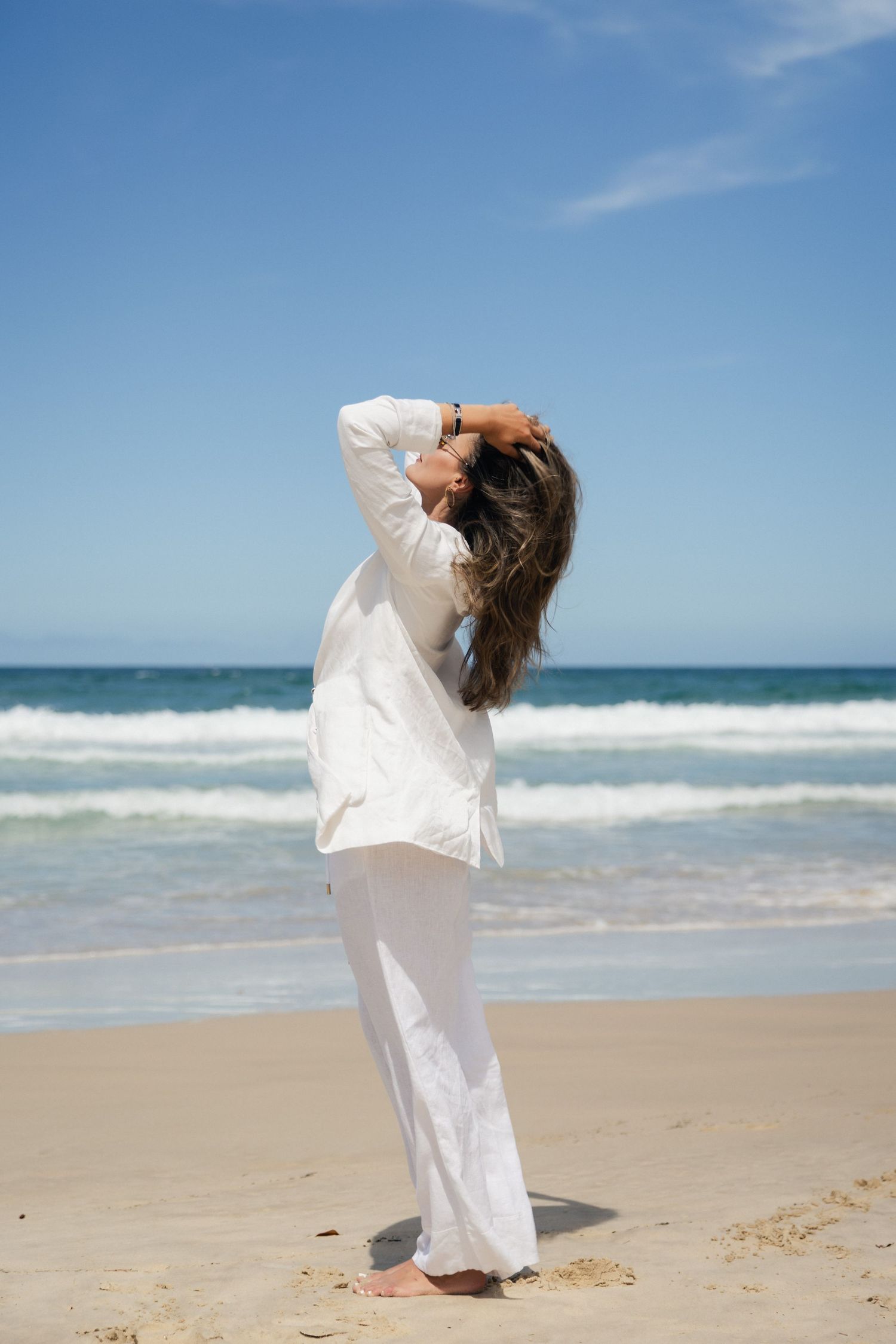 Woman in white linen outfit standing barefoot on sandy beach with ocean waves and blue sky behind her.