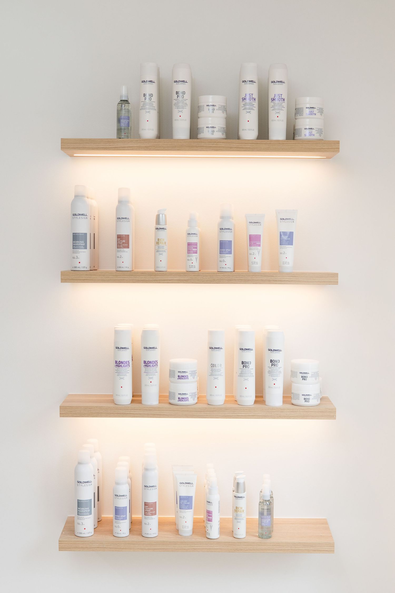 Four illuminated wooden shelves displaying Goldwell hair care products against a white wall in a salon.