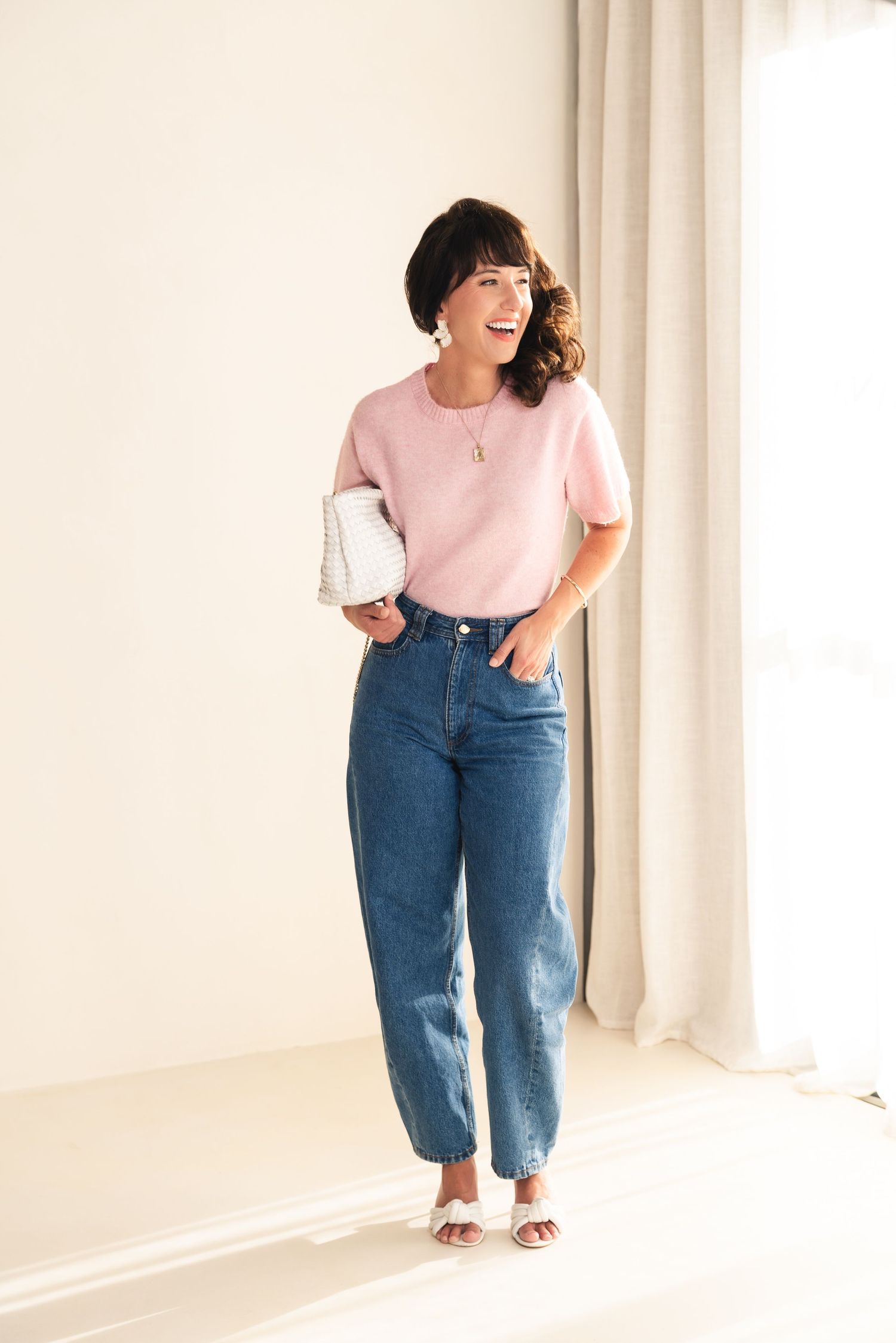 Person in light pink t-shirt and relaxed fit jeans poses against white wall in natural lighting.