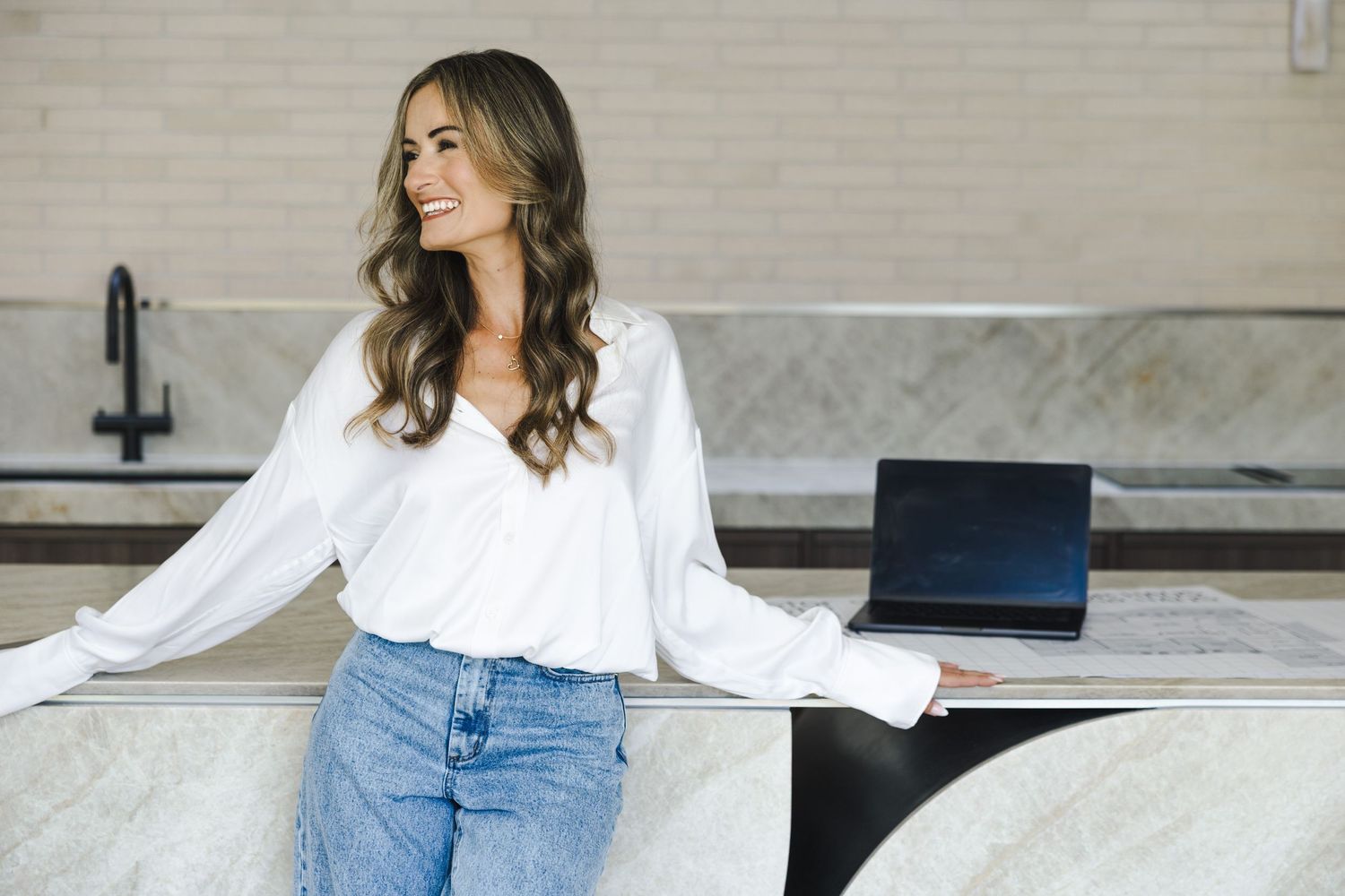 A smiling woman in a white blouse and jeans leans against a marble counter with an open laptop in a modern kitchen.