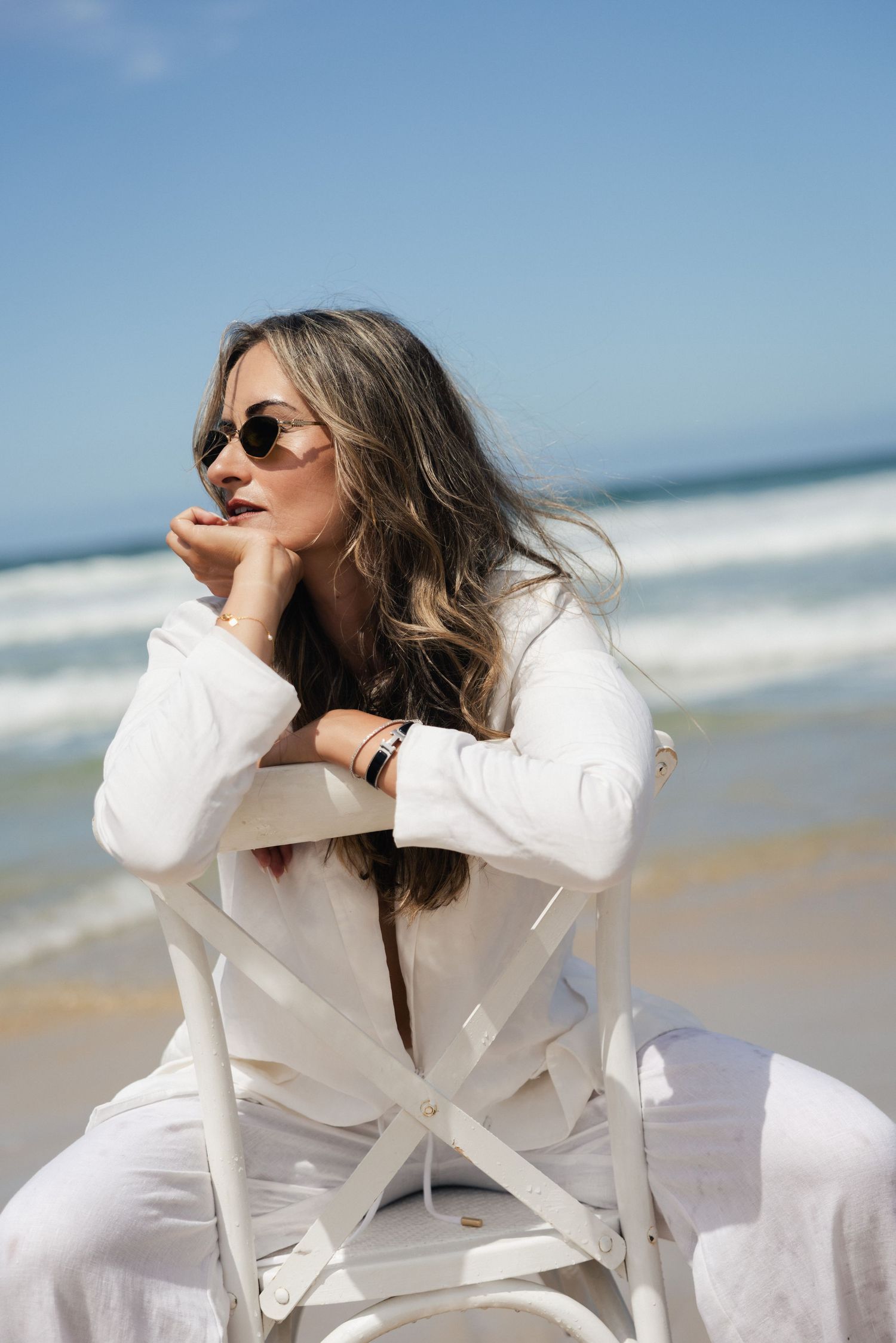Woman in white linen outfit and sunglasses sitting on white chair at beach.