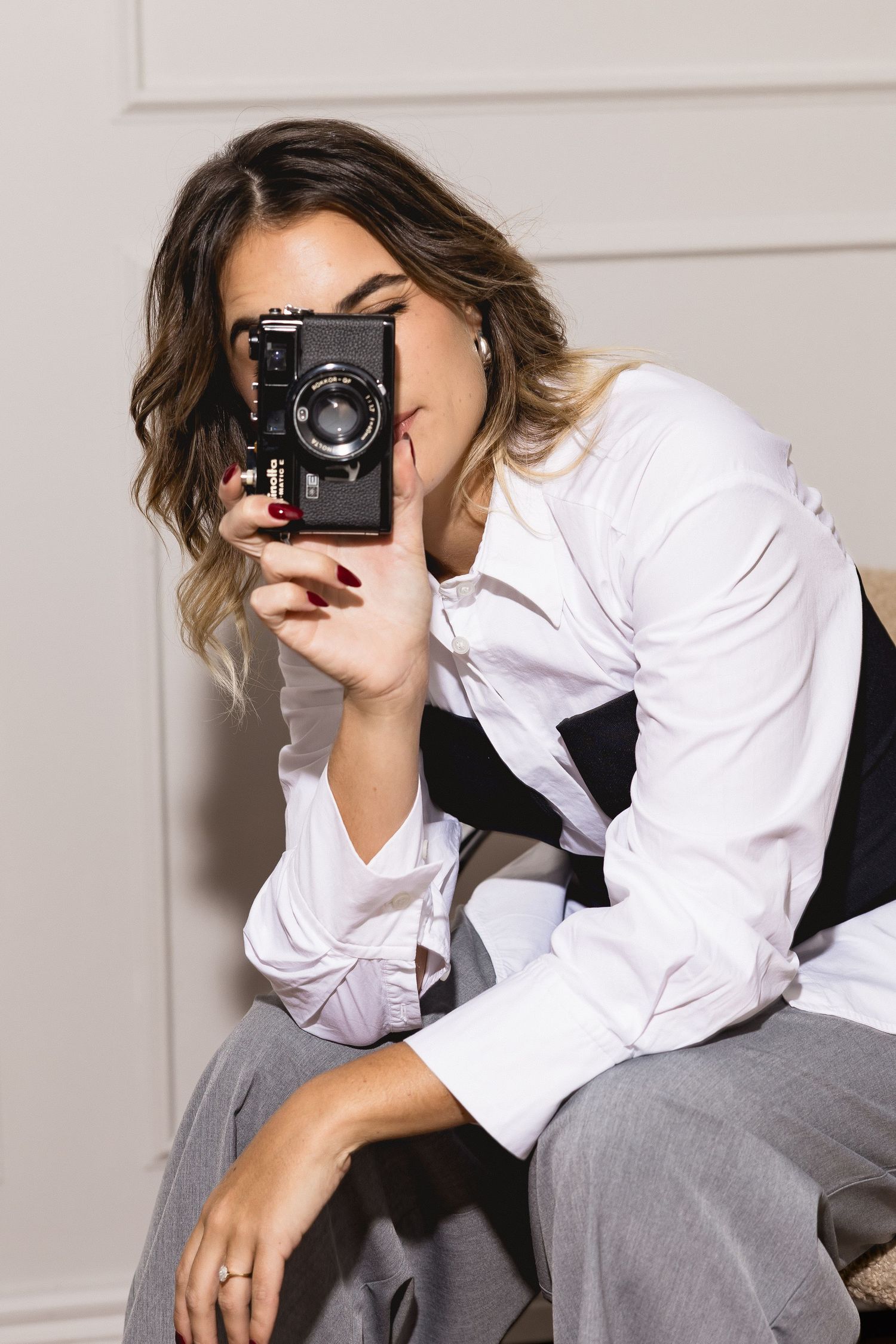 Woman in white shirt holding vintage black film camera up to her eye, wearing gray trousers, red nails.