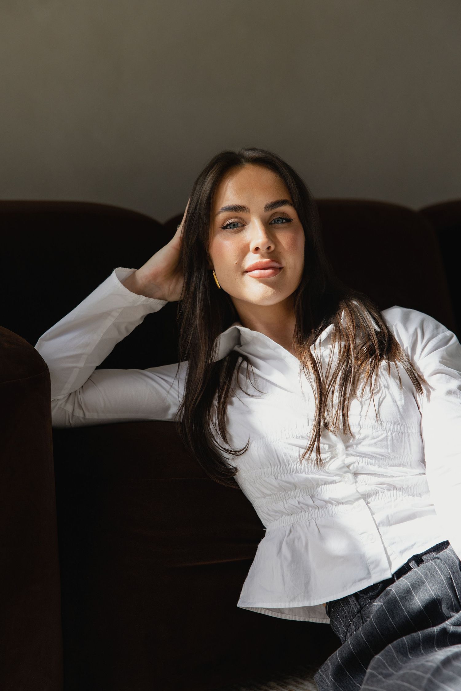 Young woman with long dark hair in white button-up shirt sitting relaxed on dark sofa, hand behind head.